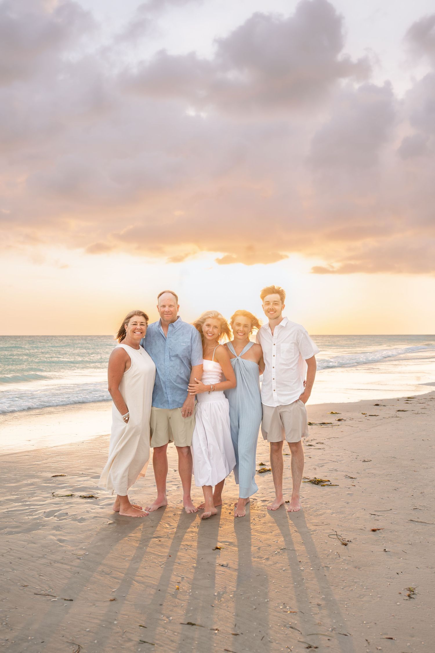 Family of five posing together on a sandy beach at sunset with colorful pastel sky and ocean waves in the background.