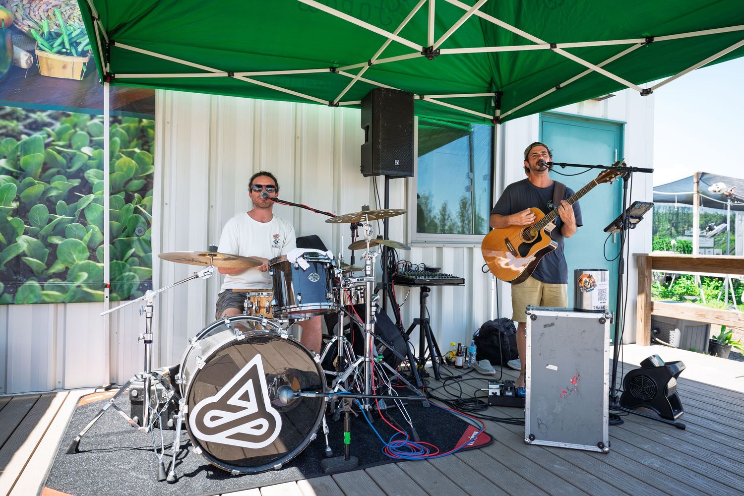 Two musicians perform outdoors on a deck stage under a green canopy, one playing drums and one playing acoustic guitar.