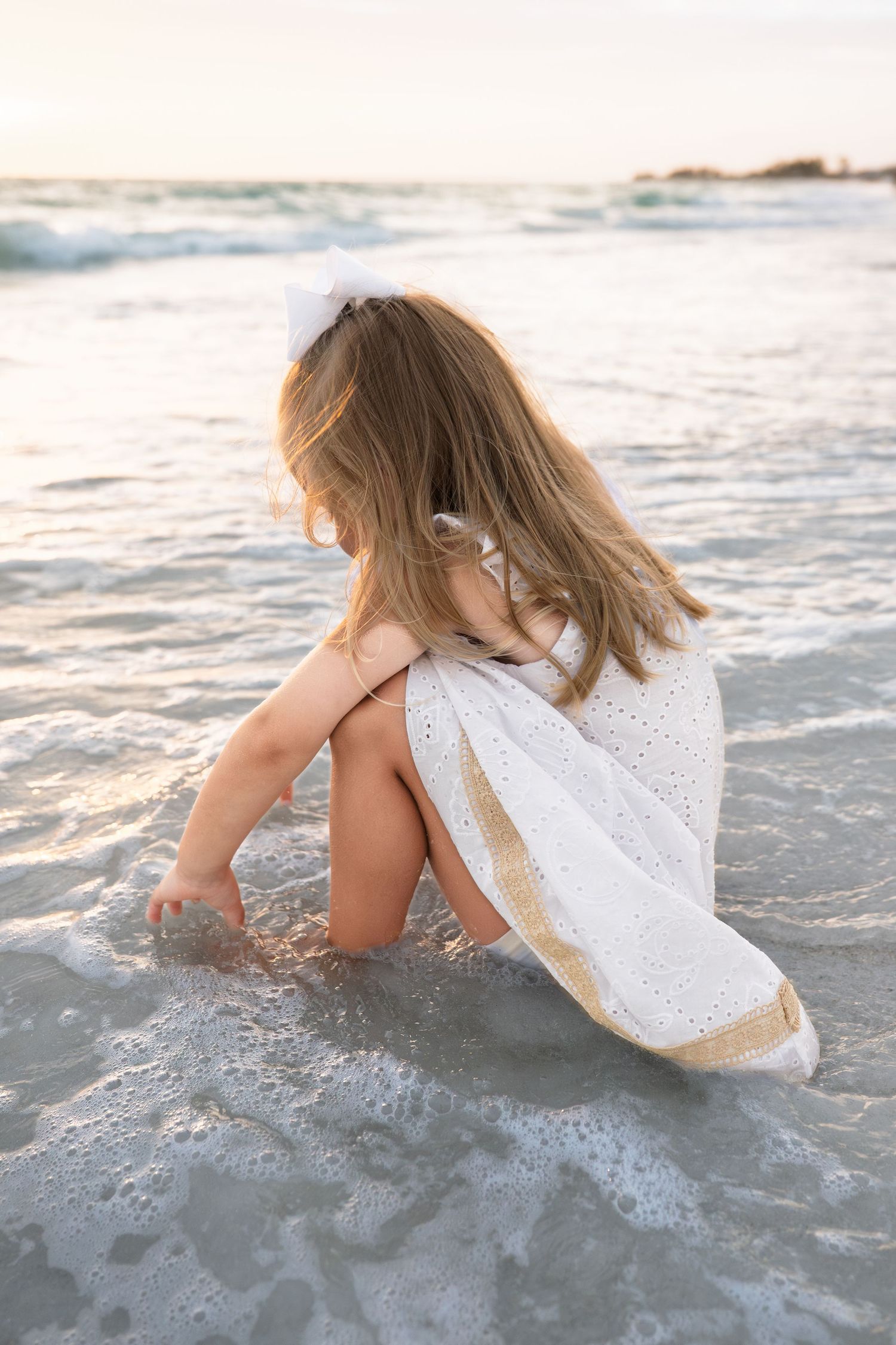 Young girl in white dress sitting at ocean's edge, playing in gentle waves at sunset on a sandy beach.