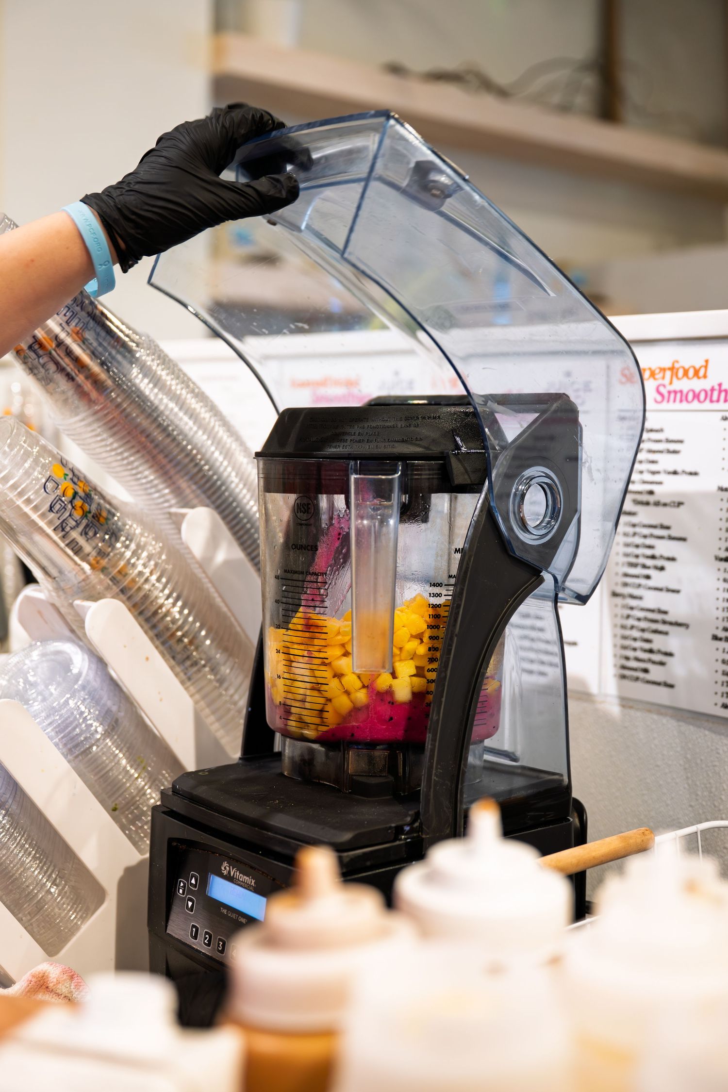 Person in black gloves operating a commercial blender with colorful fruits at a smoothie bar counter.