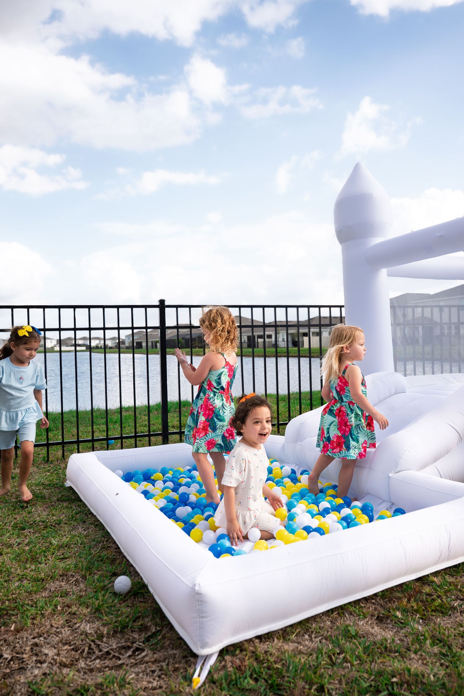 Children play in an inflatable ball pit with white unicorn design set up outdoors near a waterfront fence.