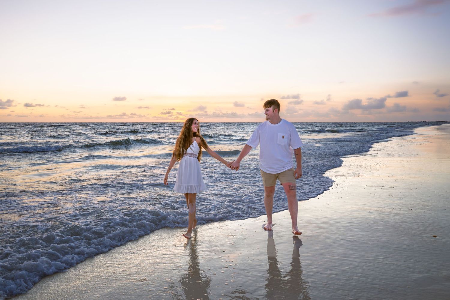 Couple holding hands walking on a sandy beach at sunset with gentle waves in the background.