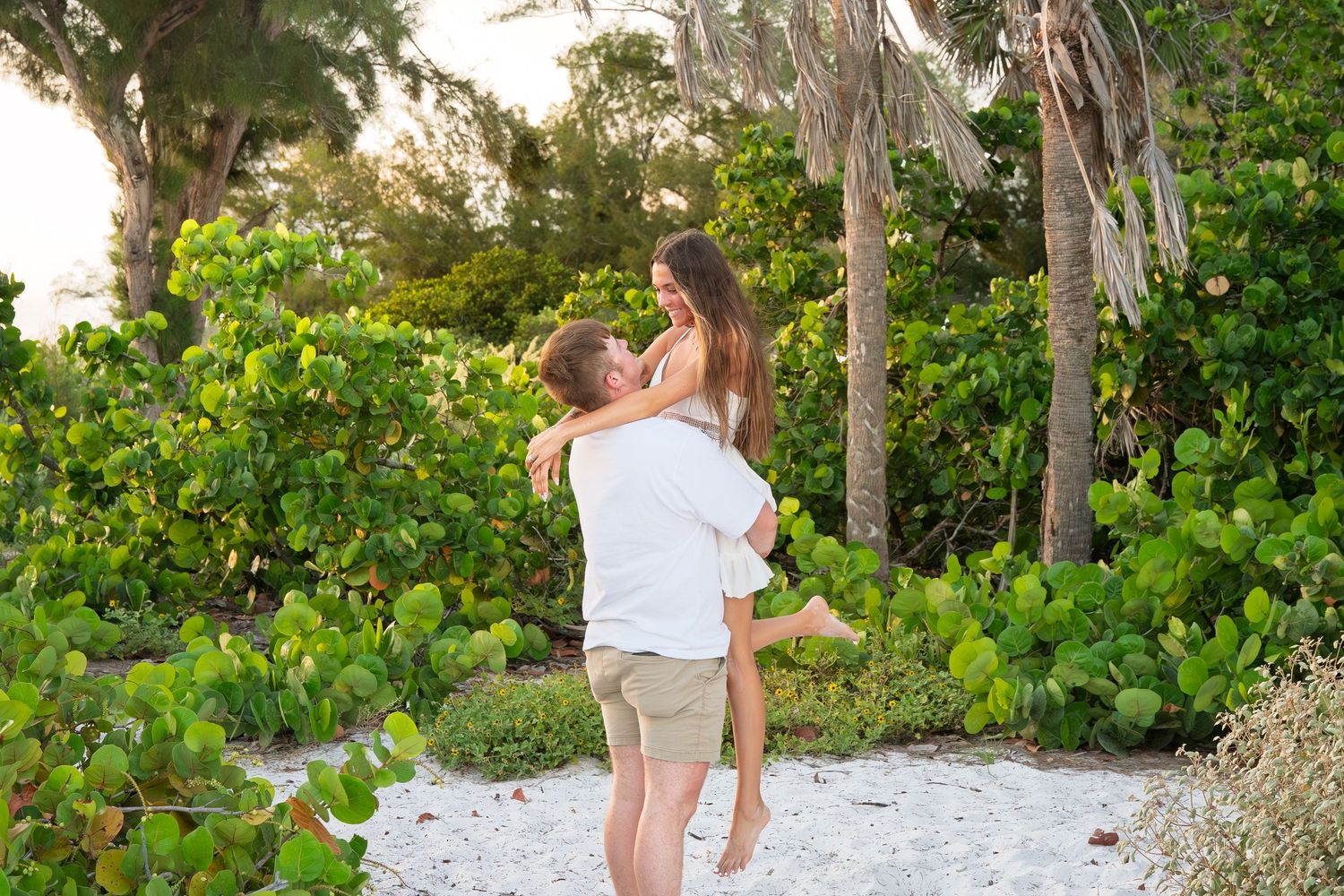 Couple embracing on a tropical beach path surrounded by lush green trees and palms at sunset.