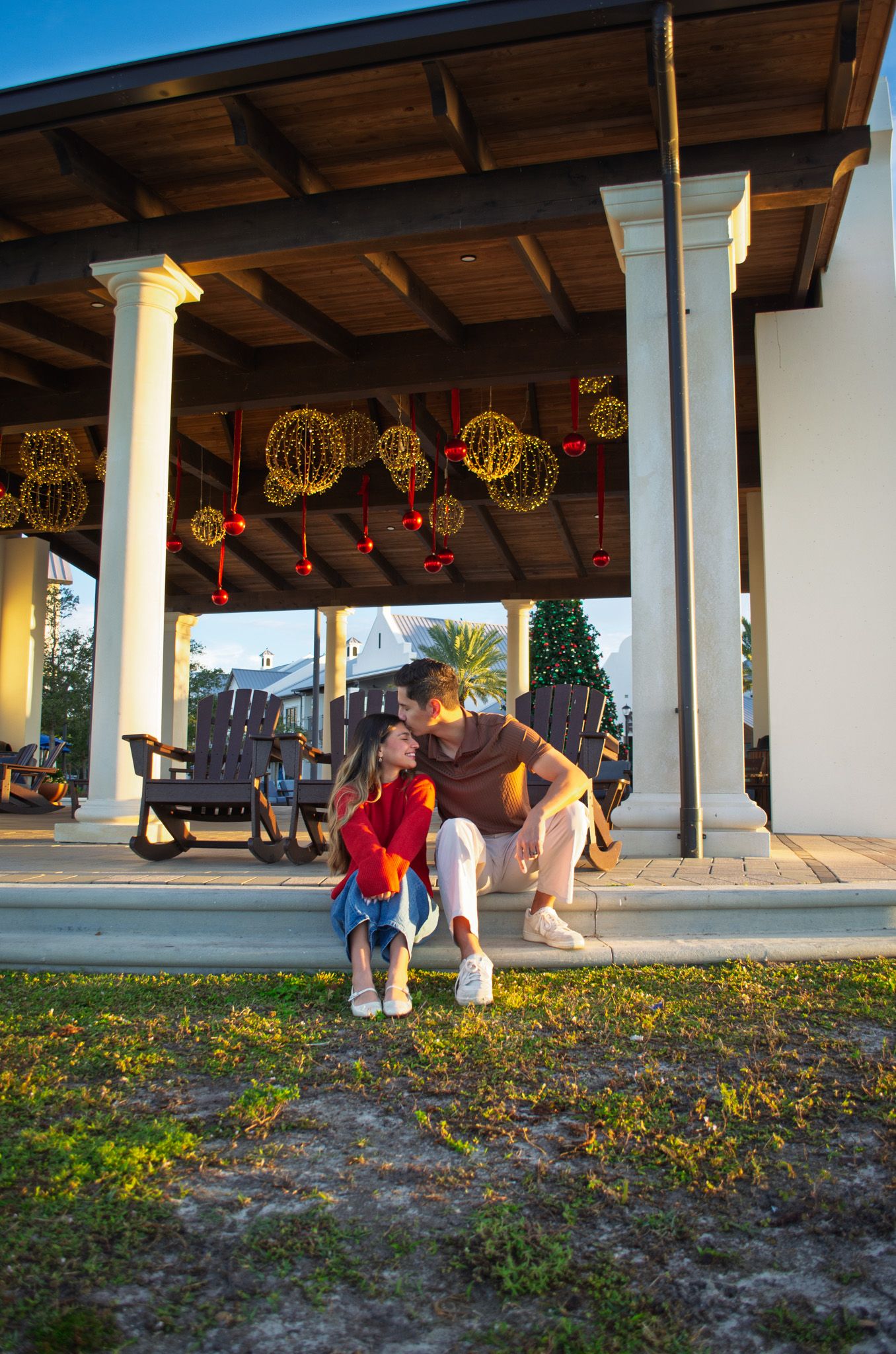 People walk under a covered walkway decorated with illuminated holiday spheres at sunset while yellow wildflowers bloom below.