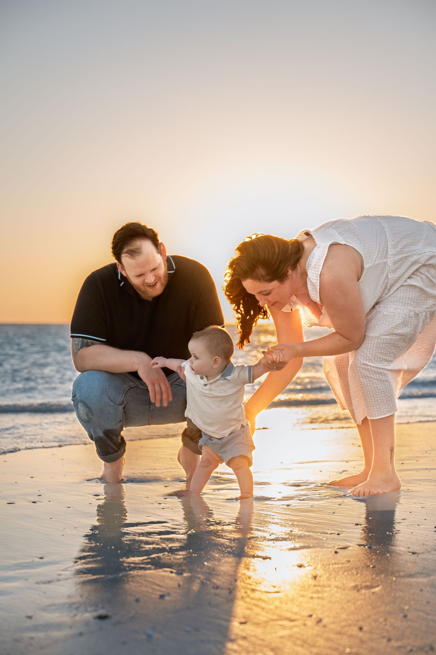 Family helping toddler take first steps on sandy beach at golden sunset, waves reflecting warm light.