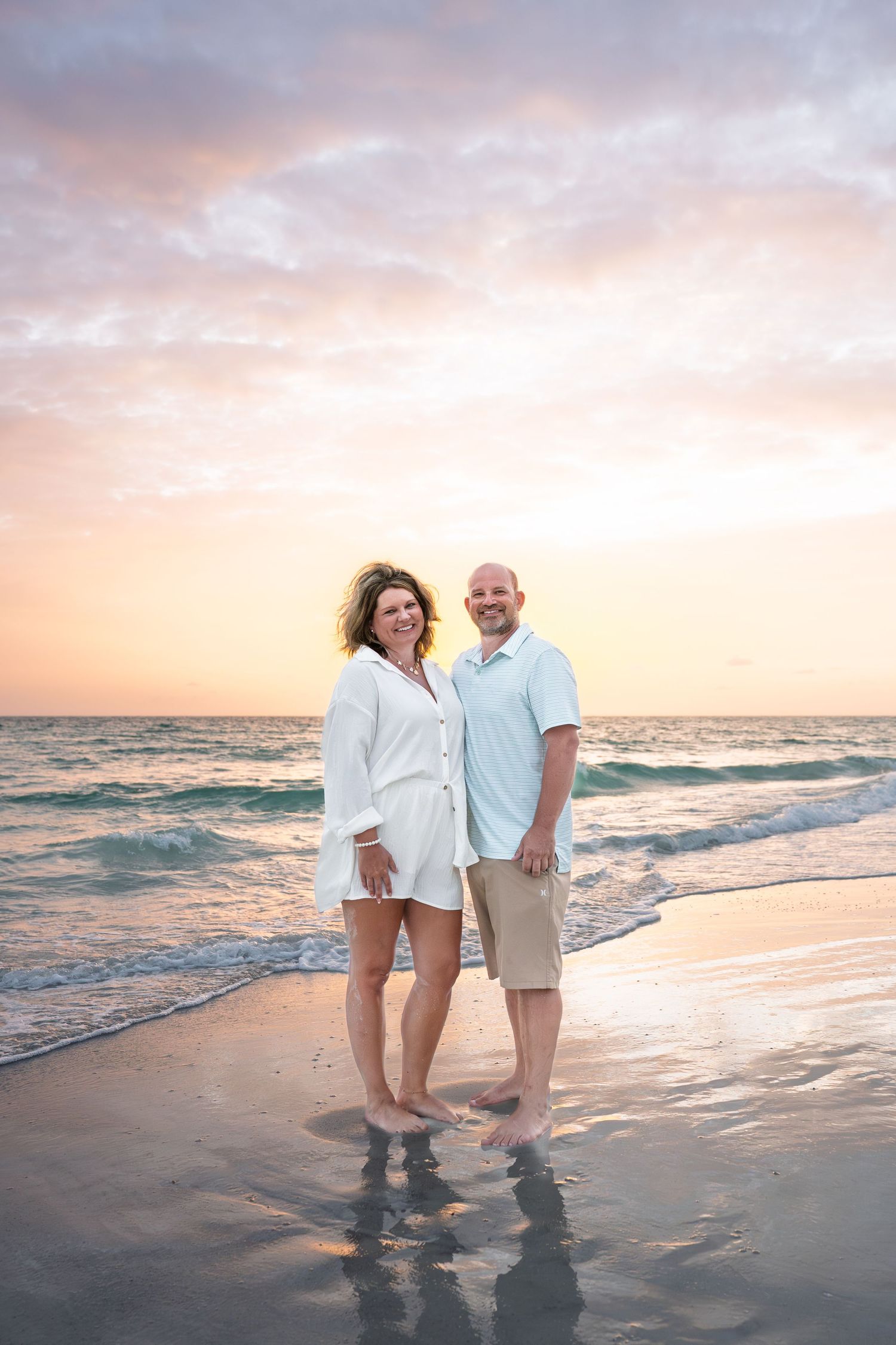Couple smiling together on a sandy beach at sunset, waves behind them, wearing casual white and blue outfits.