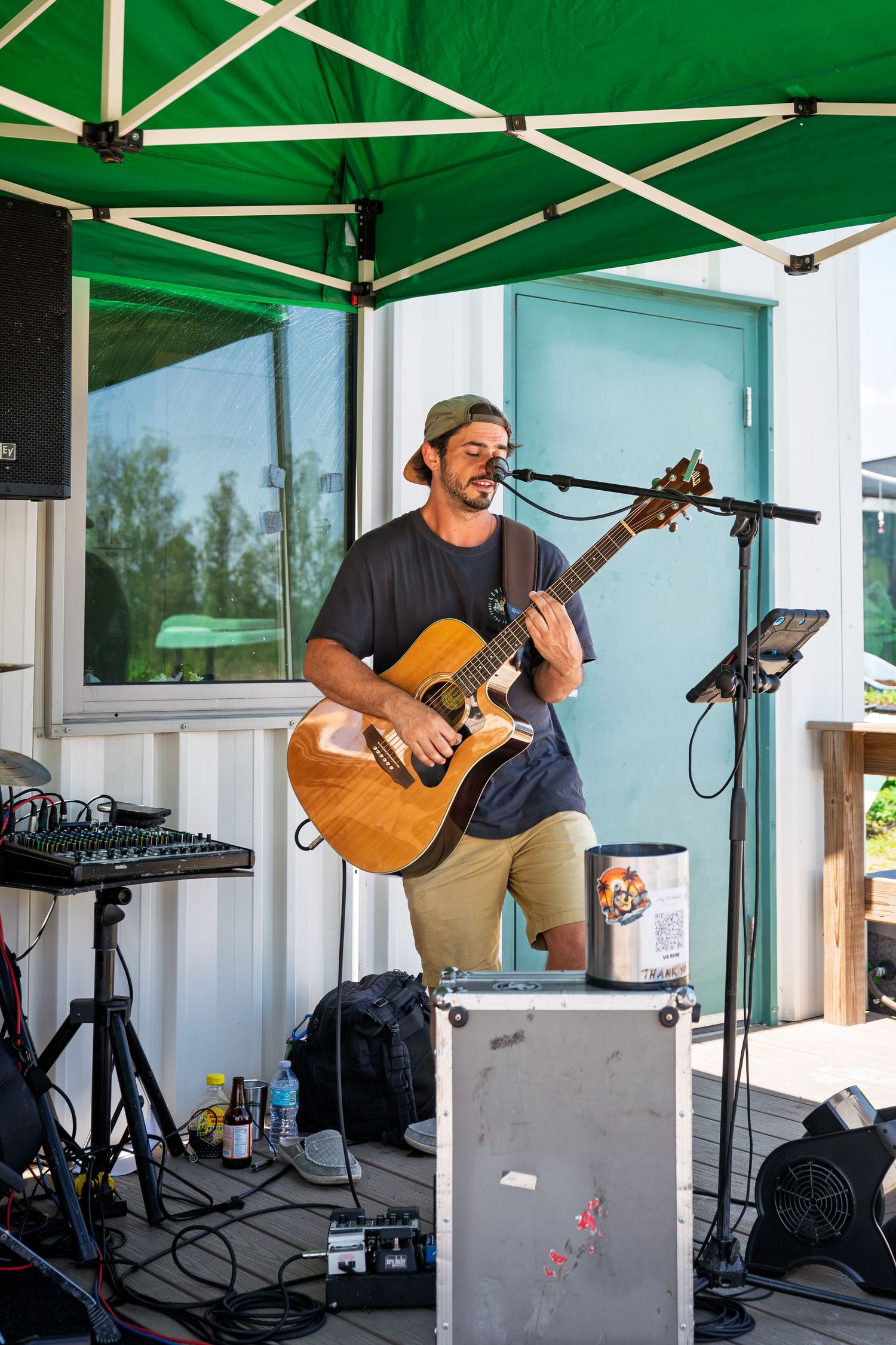 Male musician playing acoustic guitar and singing into microphone outdoors under green canopy tent at live performance.
