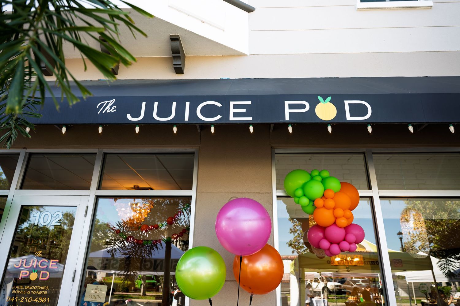 Colorful balloon clusters decorate the storefront of The Juice Pod, a fresh juice bar with a dark navy sign.