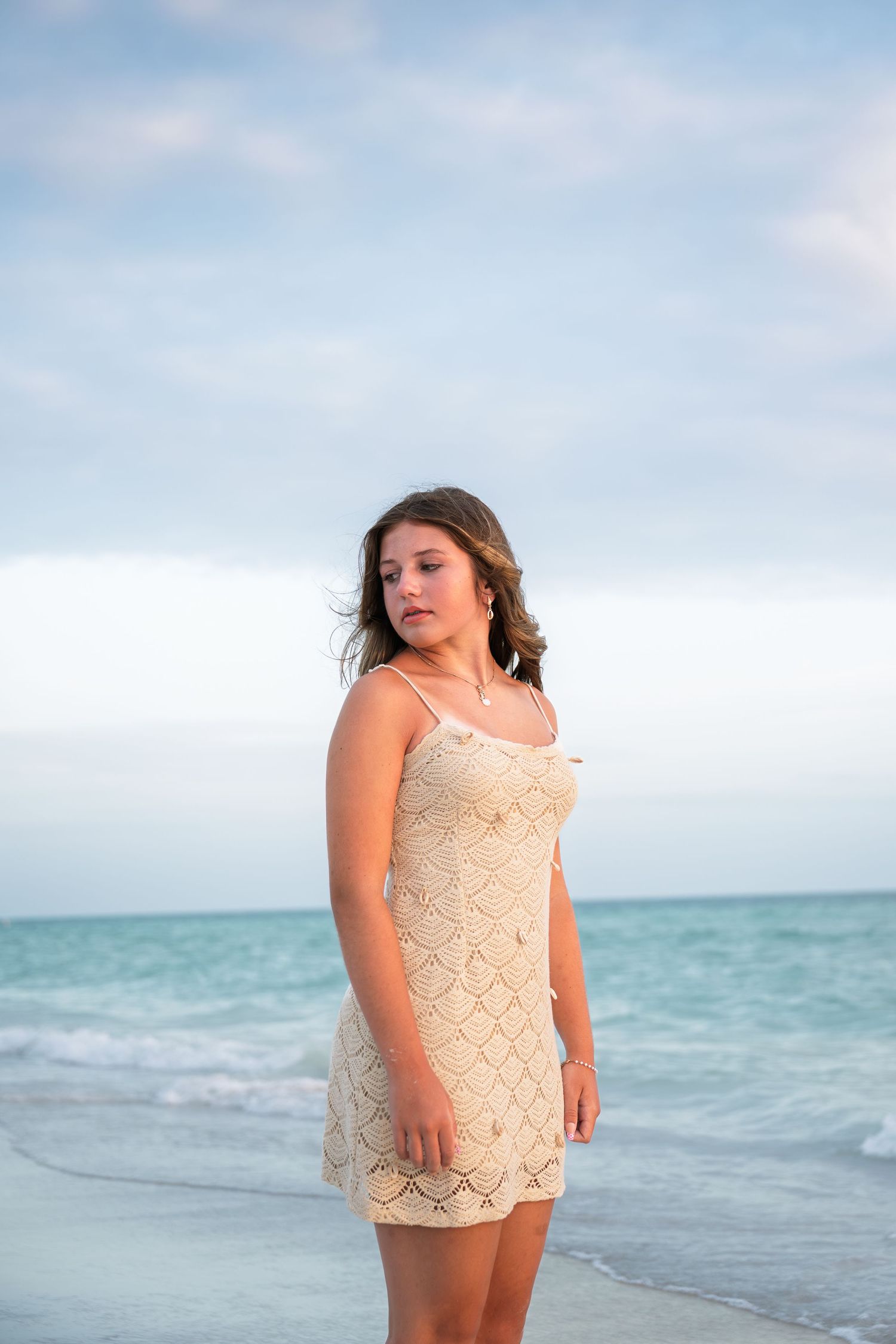 Young woman in cream lace dress standing on a beach with turquoise ocean waves in the background.