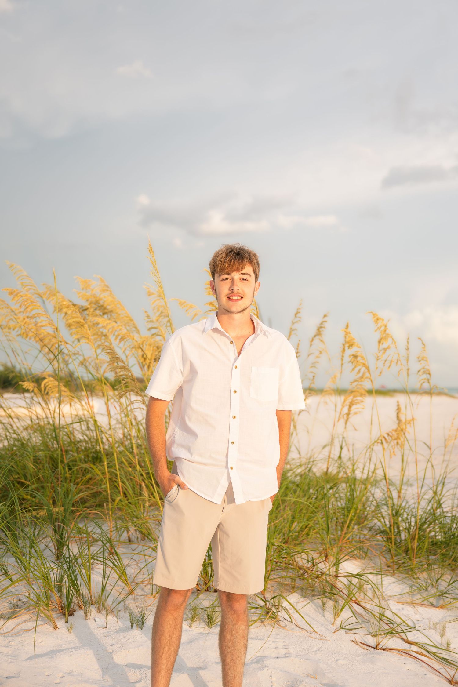 Young man in white shirt and khaki shorts standing on white sand beach with sea oats in background.