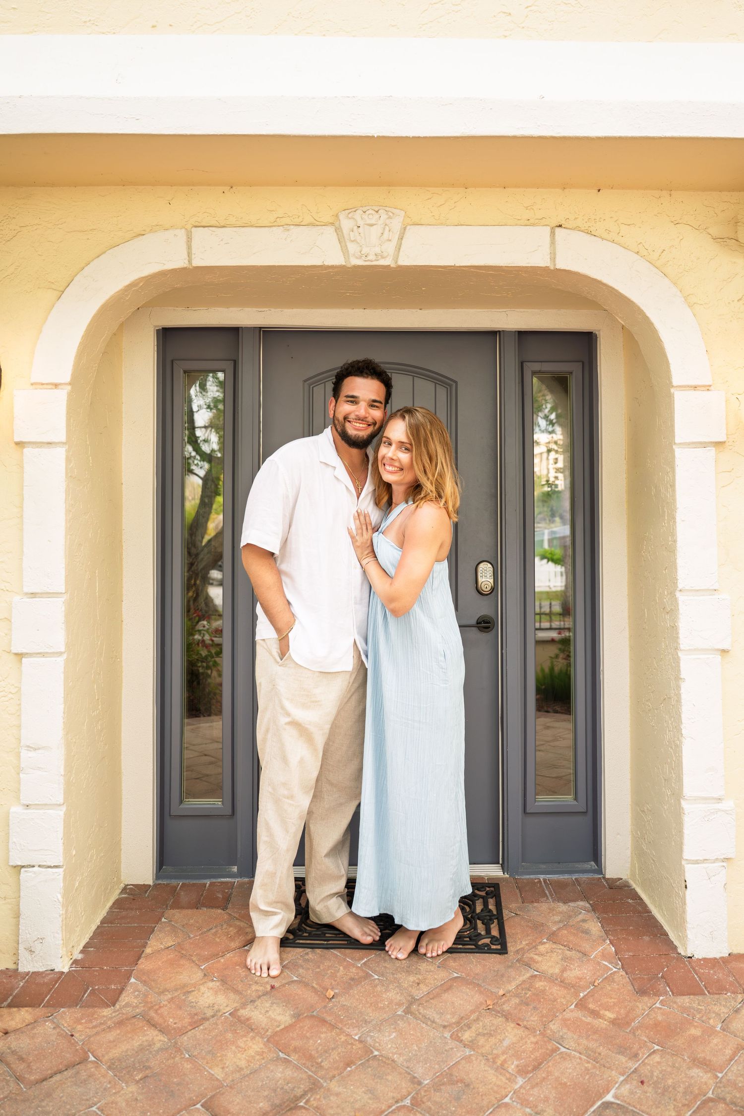 A smiling couple poses barefoot in front of a gray front door under a yellow stucco archway.