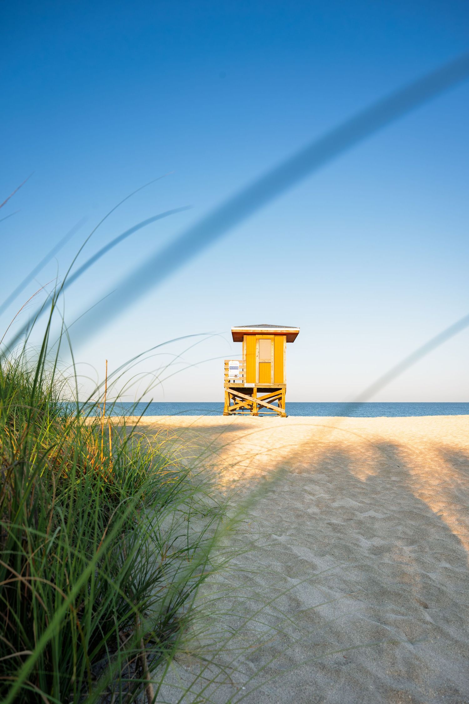 A bright yellow lifeguard tower stands on a sunny sandy beach with sea grass in the foreground and clear blue sky.