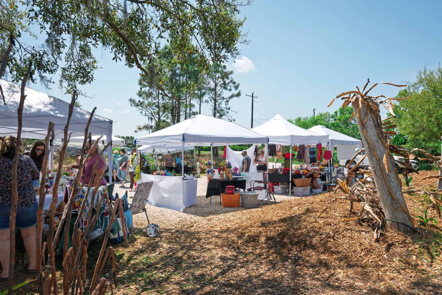 Outdoor art market with white canopy tents displaying colorful artwork among shady trees on a sunny day.