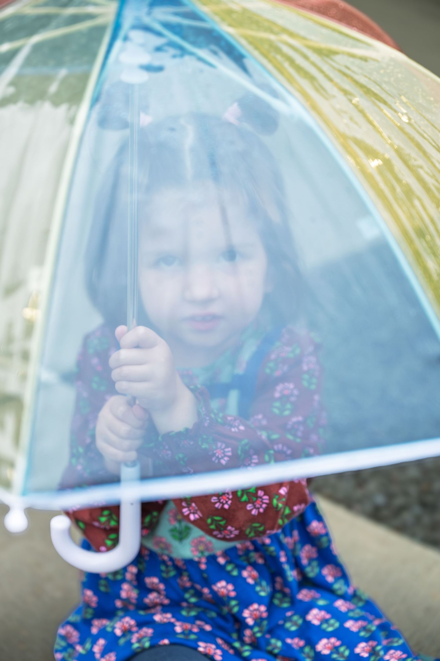Young girl in floral dress holding a colorful transparent rainbow umbrella outdoors on a rainy day.