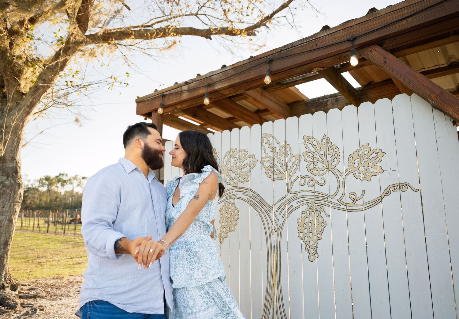 Couple sharing a romantic moment holding hands near a decorative grapevine mural on a white fence at a winery.