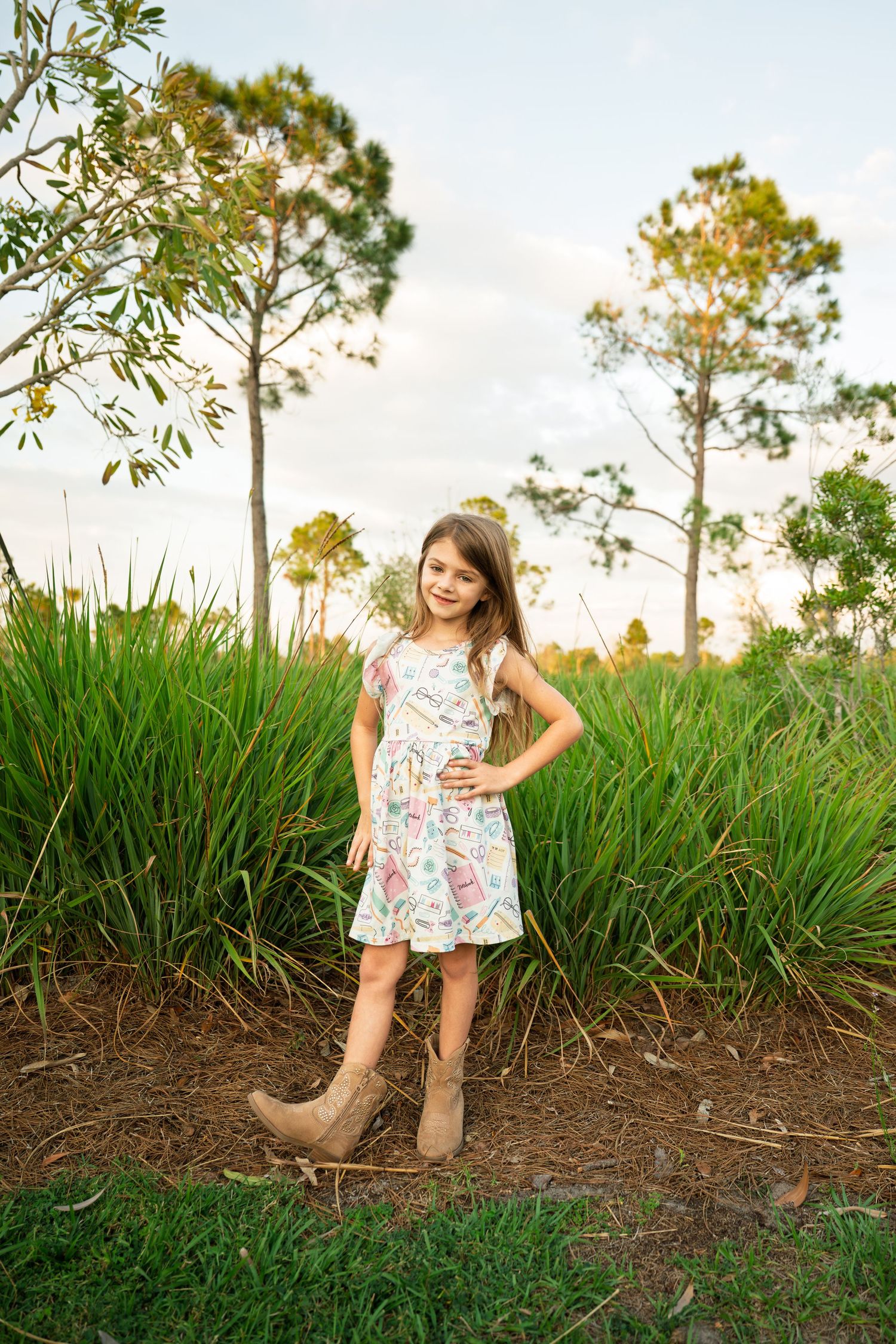Young girl in floral dress and cowboy boots posing outdoors near tall grass and pine trees at sunset.