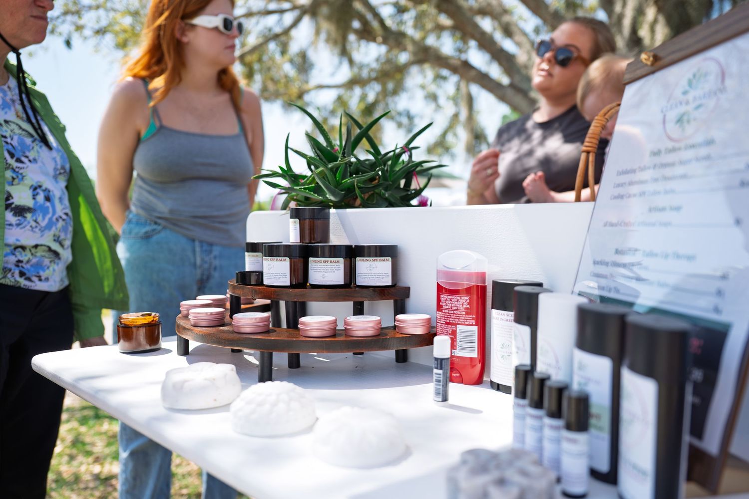 Outdoor market booth displaying artisan skincare products, candles, and succulents on a white table with shoppers browsing.