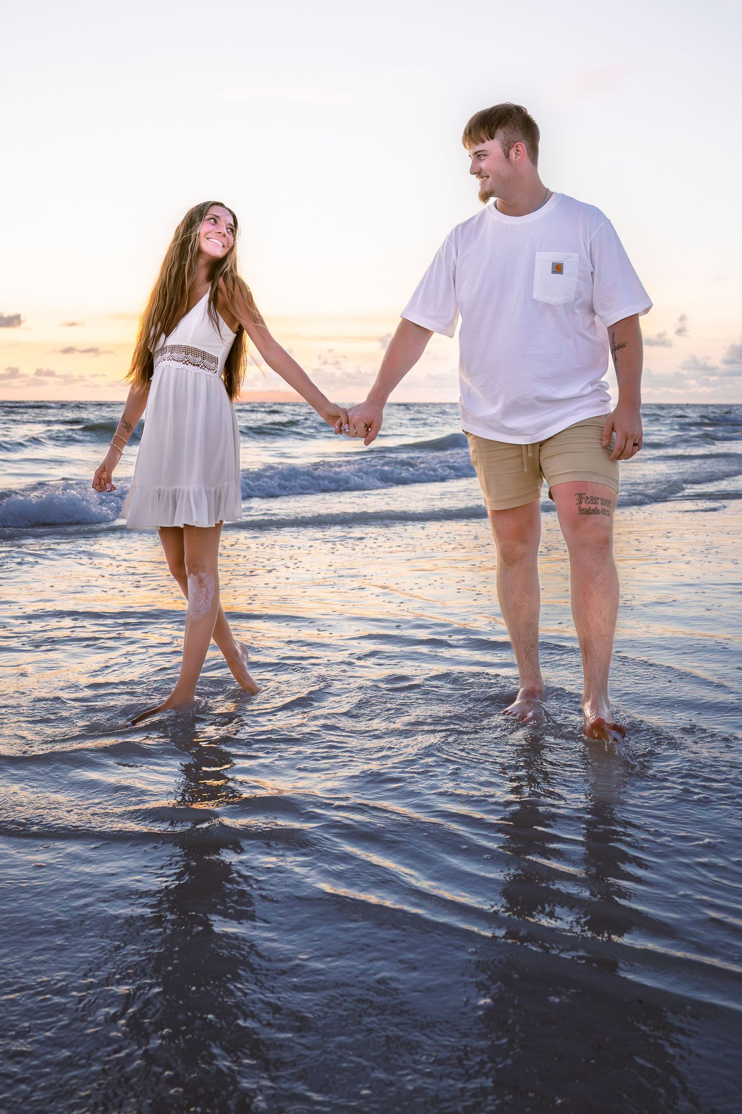 A couple holds hands walking barefoot on a beach at sunset, woman in white dress, man in white shirt and khaki shorts.