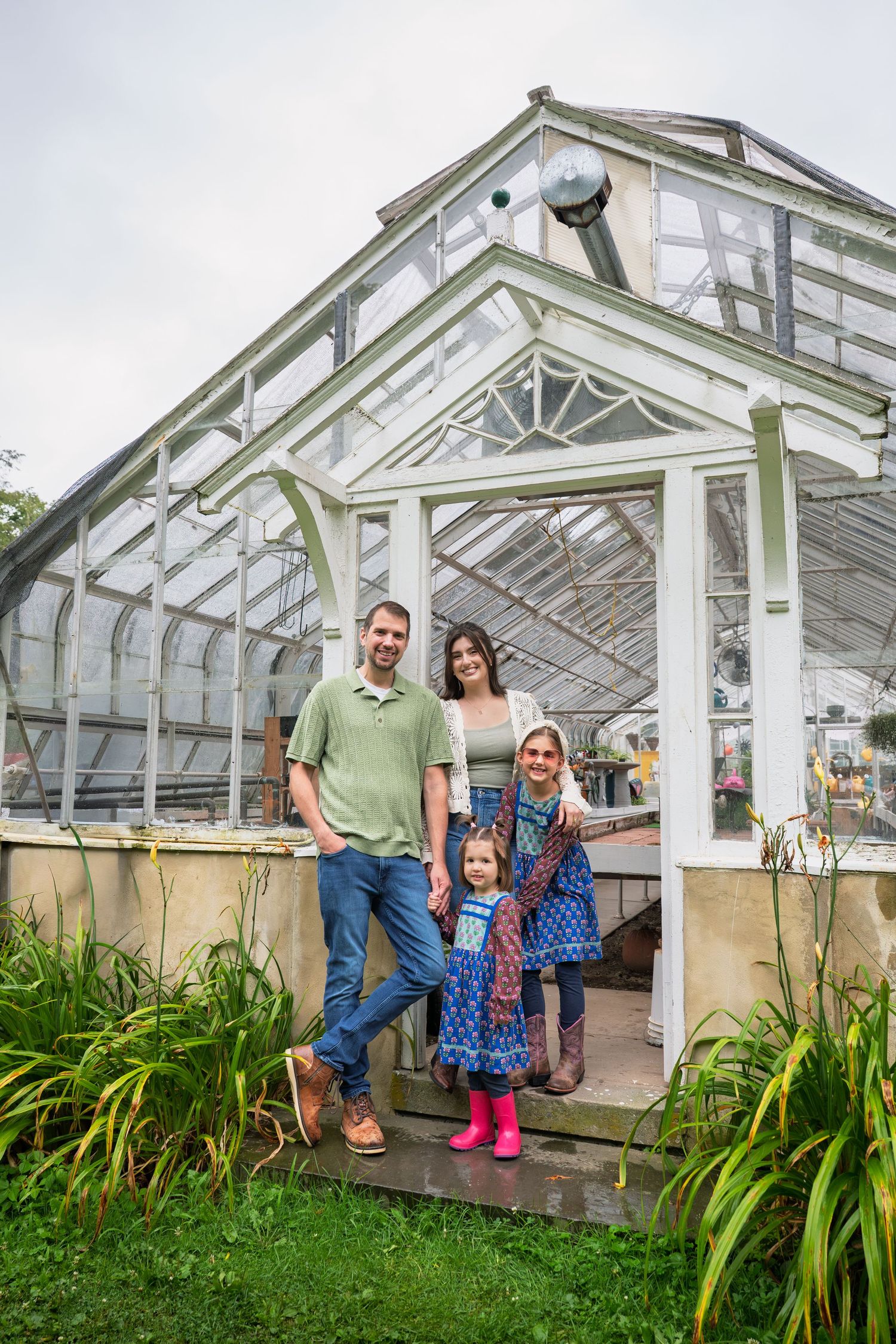 Family of three posing outside a vintage glass greenhouse surrounded by lush green plants on a cloudy day.