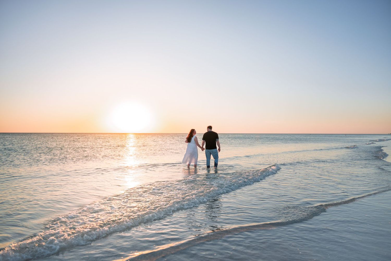 Couple holding hands on a sandy beach at sunset, waves gently washing over their feet with a golden horizon.