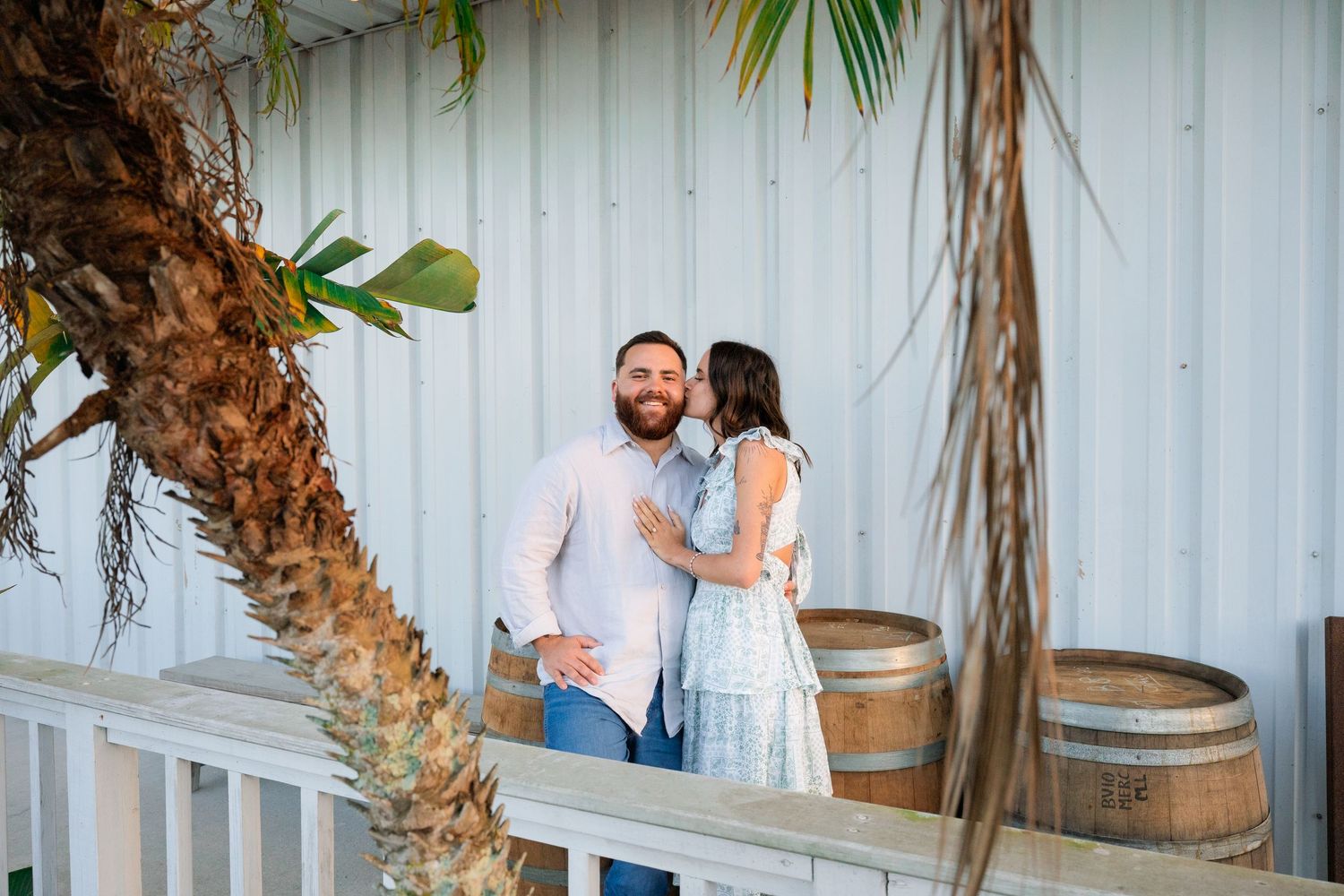 A couple embraces near rustic wooden barrels and palm trees against a white wall.