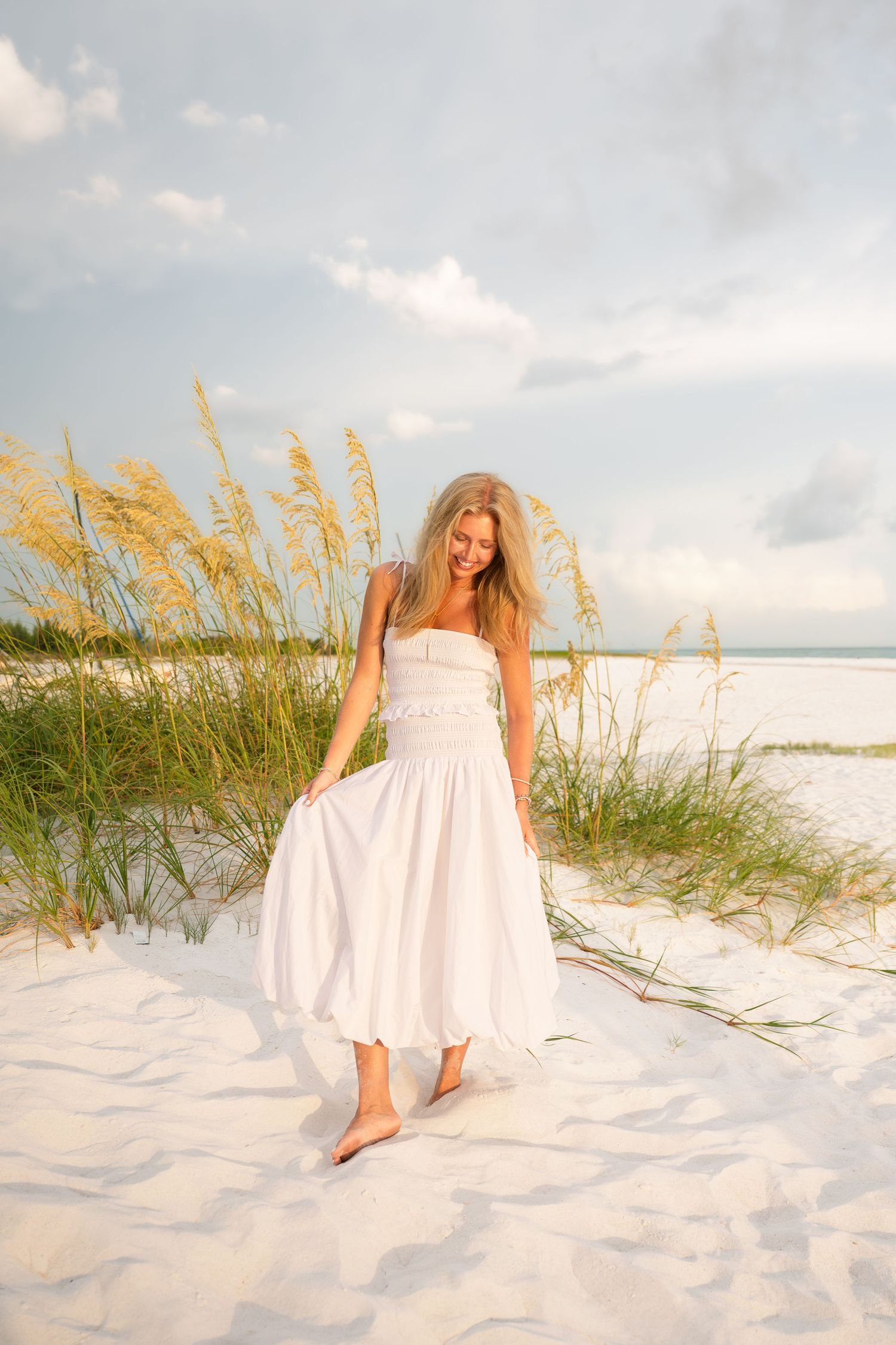 Woman in white sundress walking barefoot on white sand beach dunes with sea oats and cloudy sky.