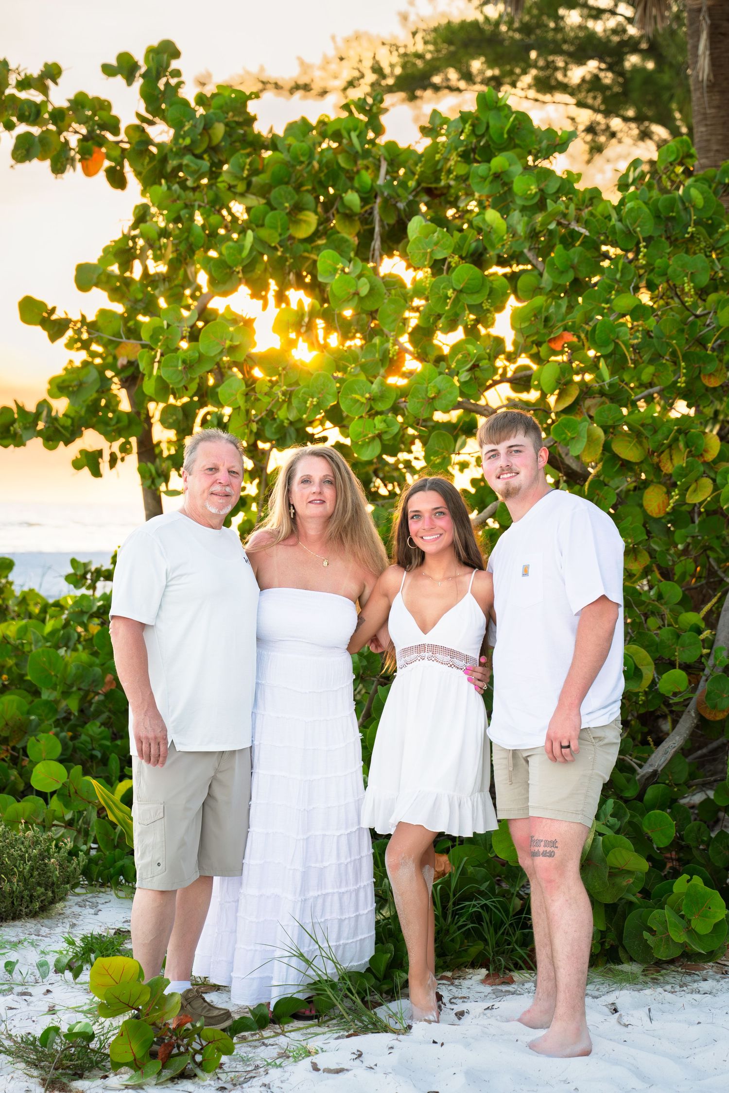 Family of four posing in white outfits on sandy beach at golden sunset with lush green trees behind them.