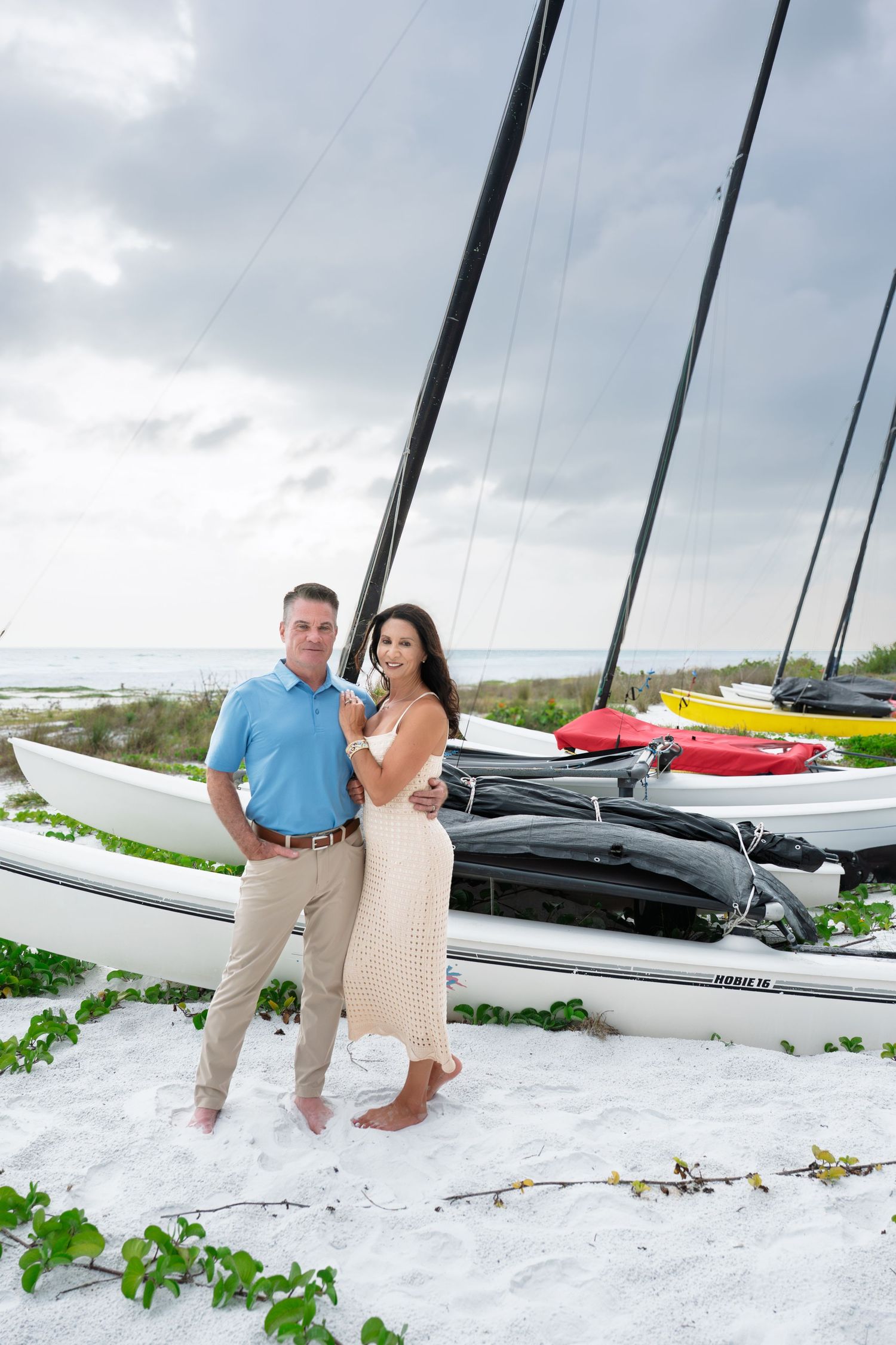 Couple posing together on white sand beach beside colorful sailboats and catamarans under cloudy skies.
