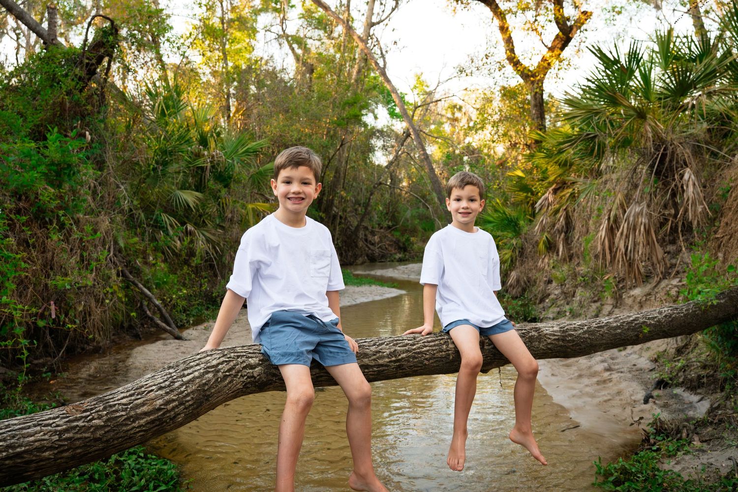 Two smiling boys in white shirts sit on a fallen tree over a sandy creek surrounded by lush green forest.