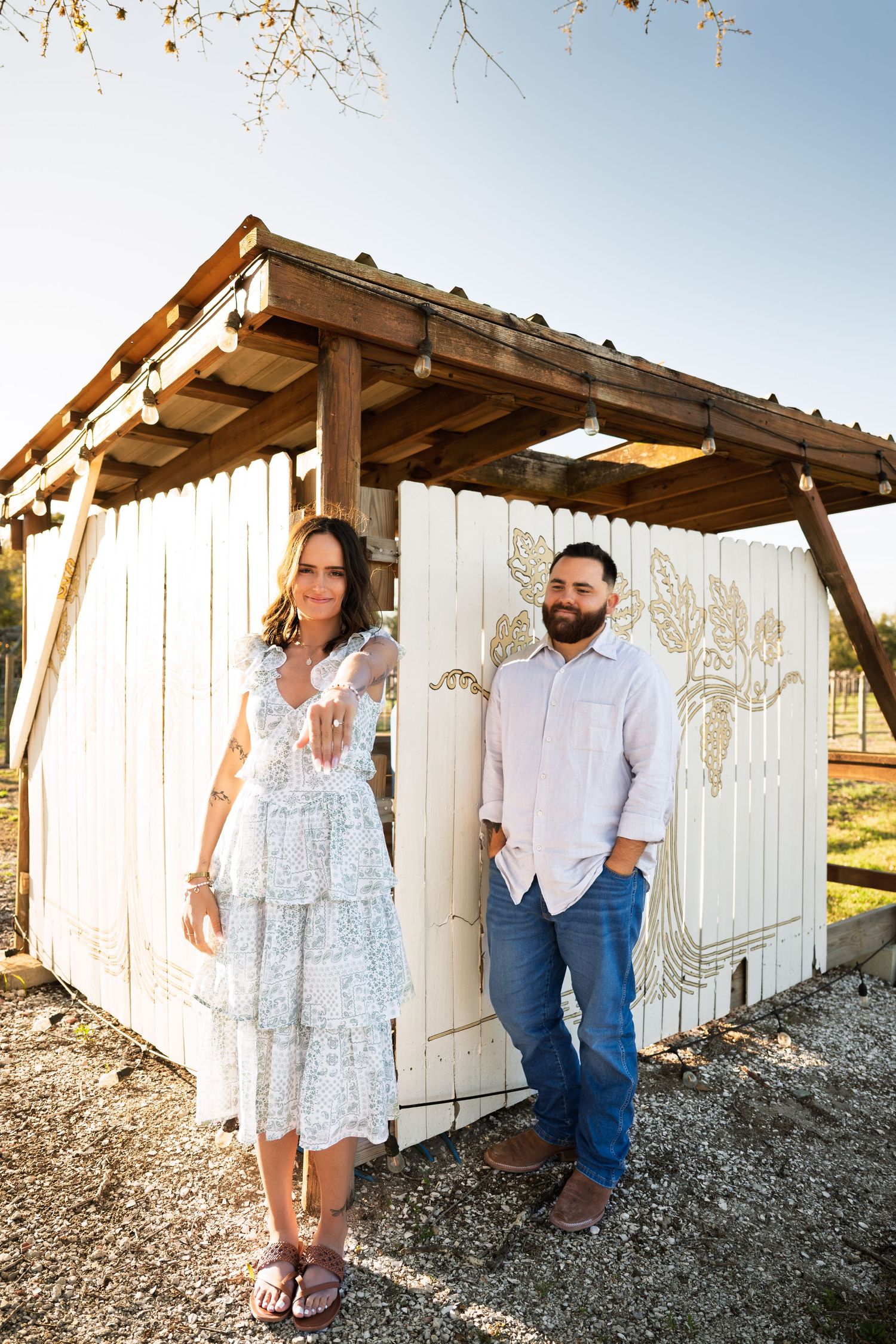 Couple in casual attire poses together against rustic white wooden wall under wooden pergola in outdoor setting.