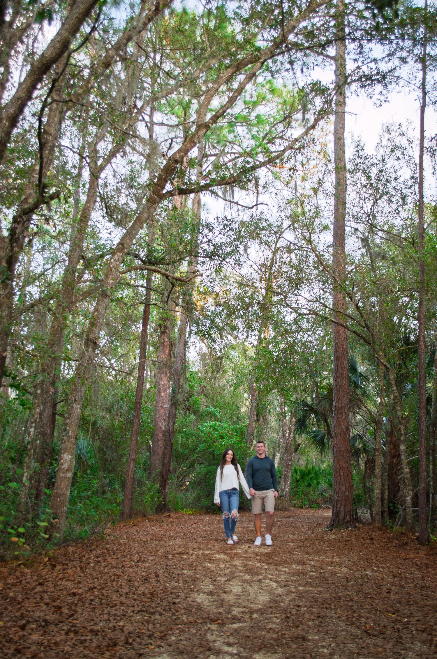 A couple walks down a tree-lined trail covered in pine needles.