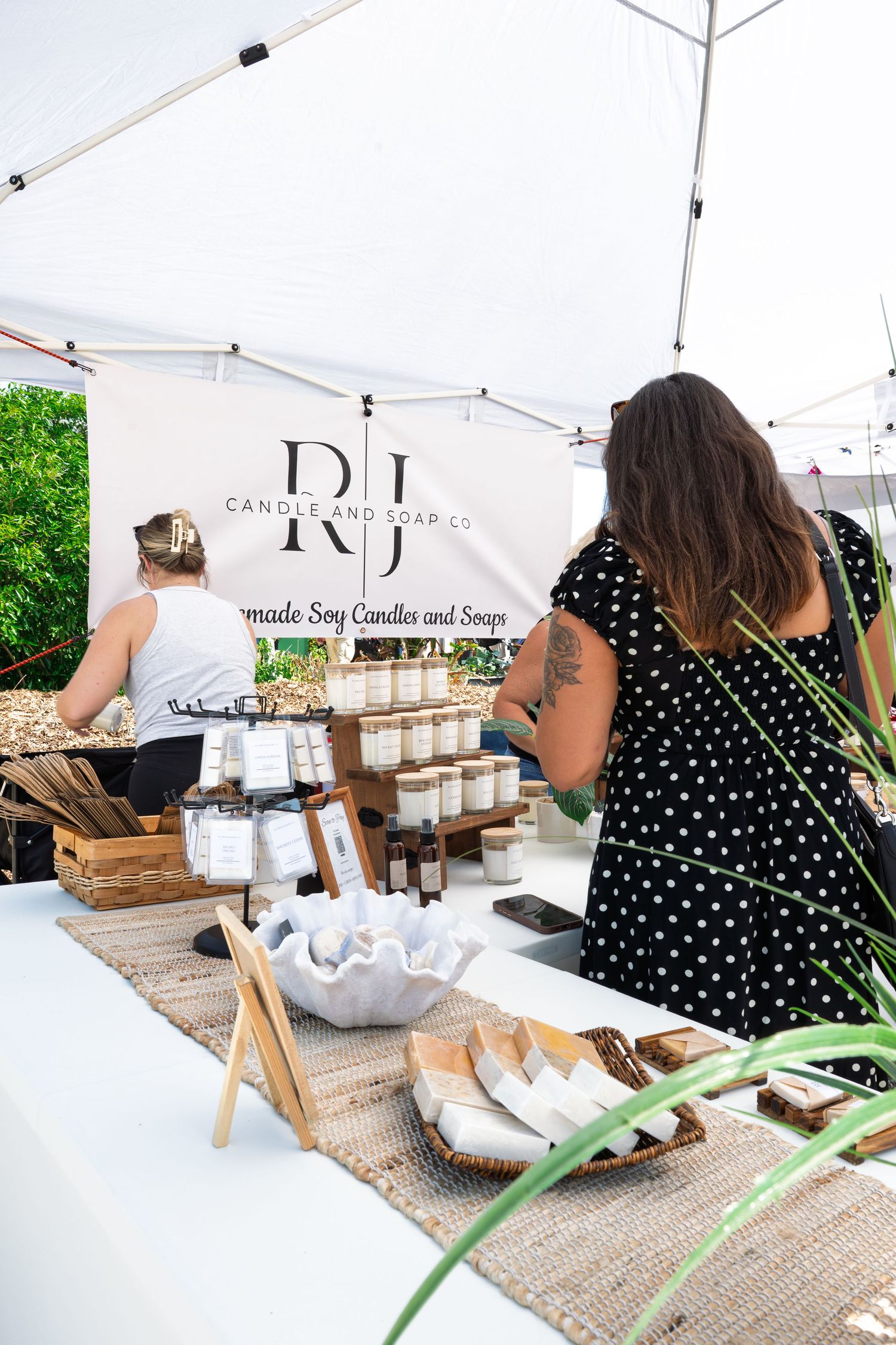 Woman shopping at RJ Candle and Soap Co. booth with handmade soy candles and soaps displayed at outdoor market.