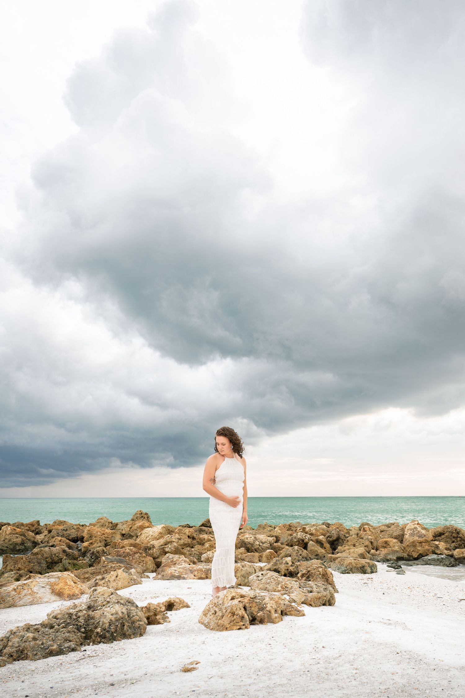 Woman in white dress standing on rocky beach under dramatic stormy sky with turquoise ocean backdrop.