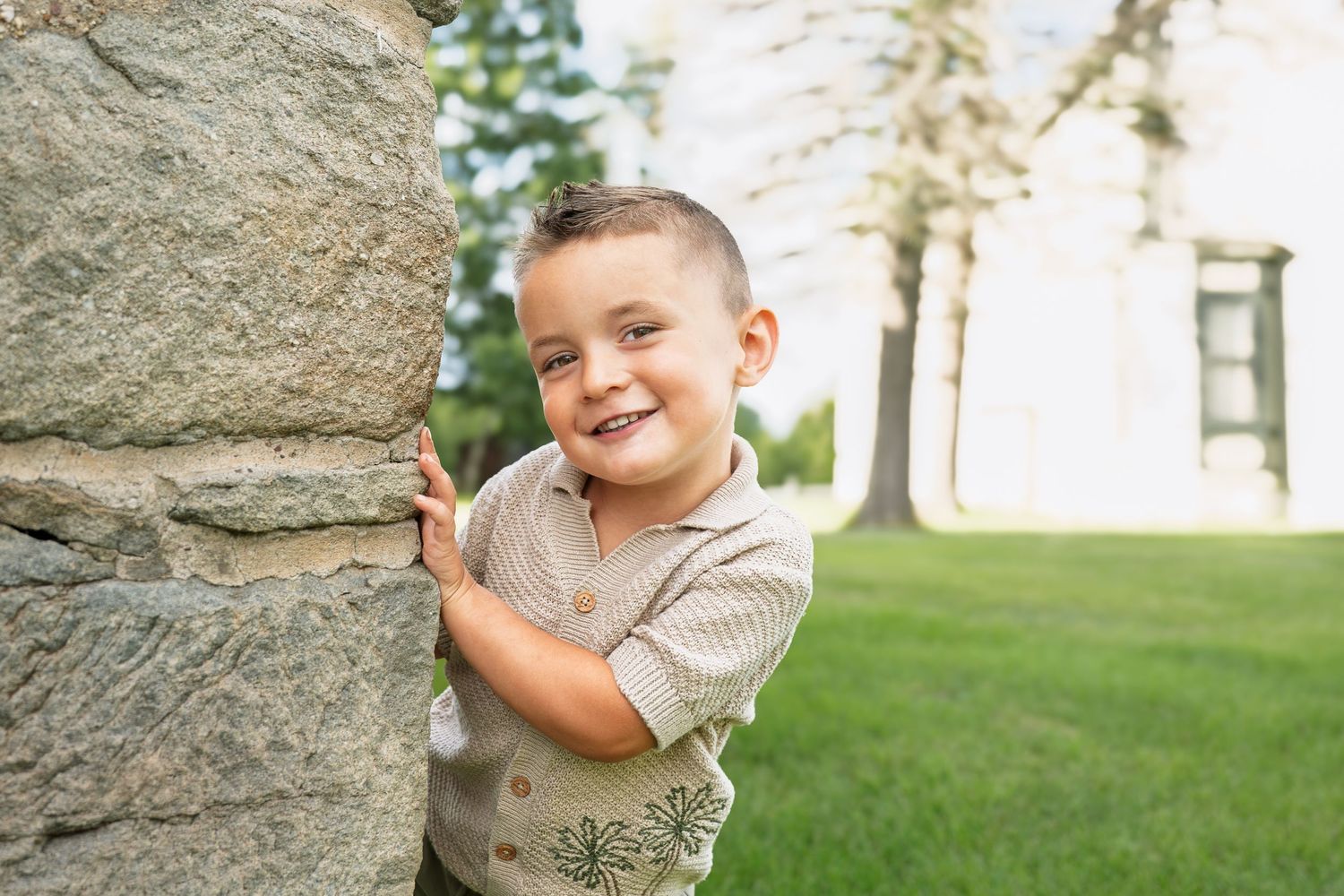 Smiling young boy peeking around a stone wall in a lush green park setting.