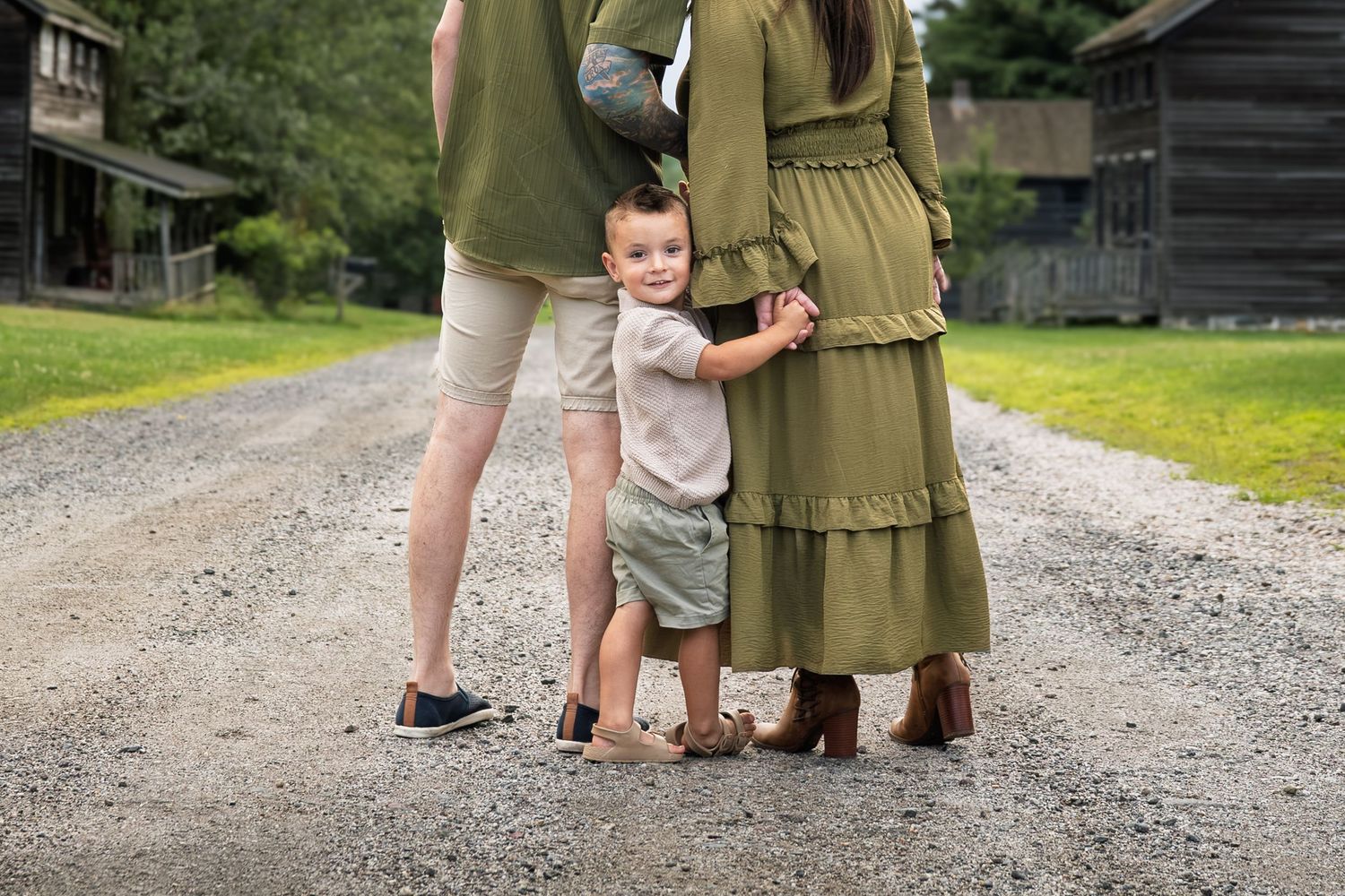 Family in matching green outfits standing on gravel path with toddler boy between parents near rustic barn.