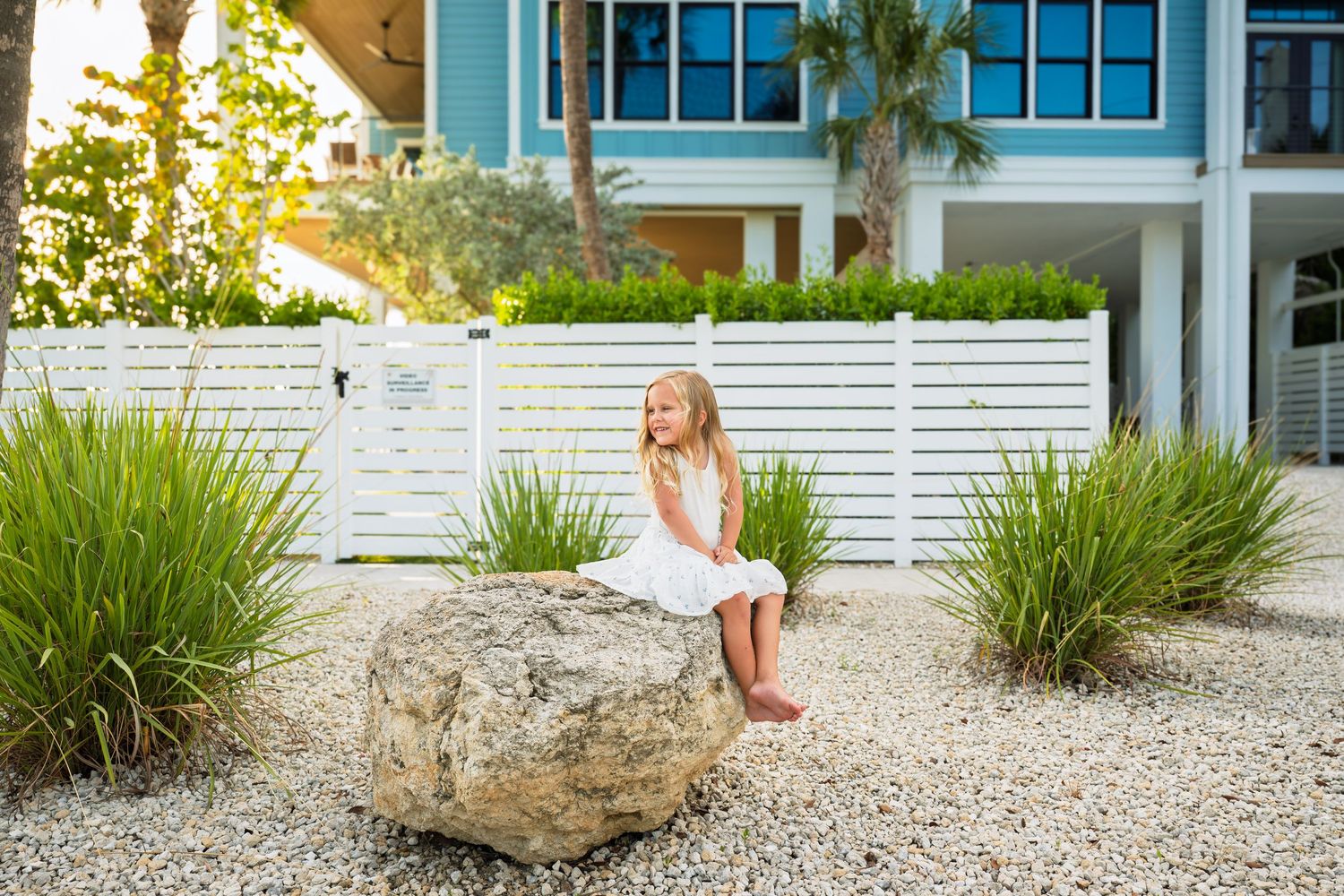 Young girl in white dress sitting on a large rock in a coastal home garden with white fence and tropical plants.