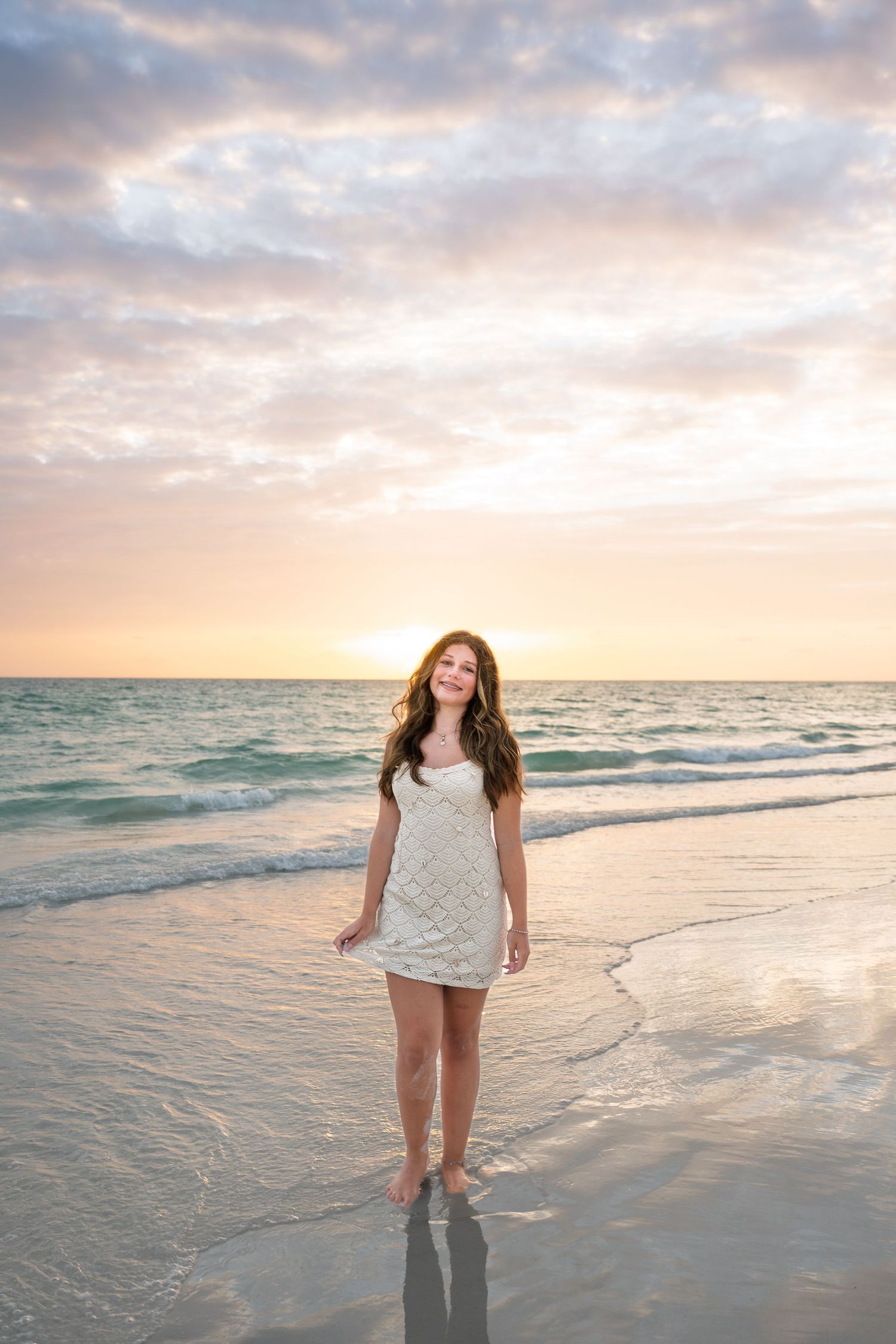 Young woman in white dress standing barefoot on a sandy beach at sunset with colorful sky and ocean waves.