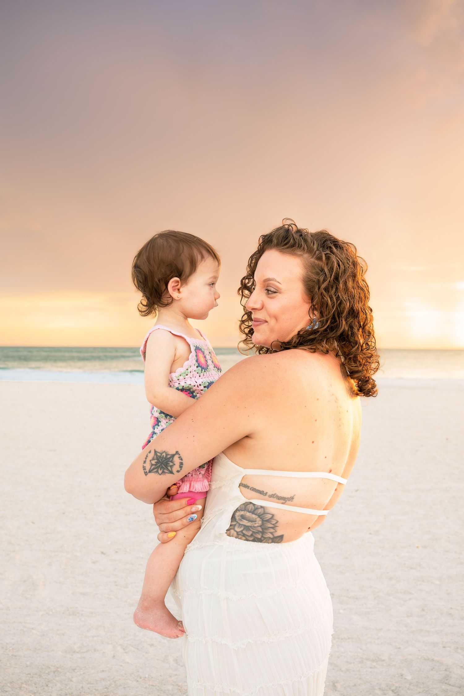 A tattooed mother holds her baby girl on a sandy beach at sunset, both wearing summer outfits.