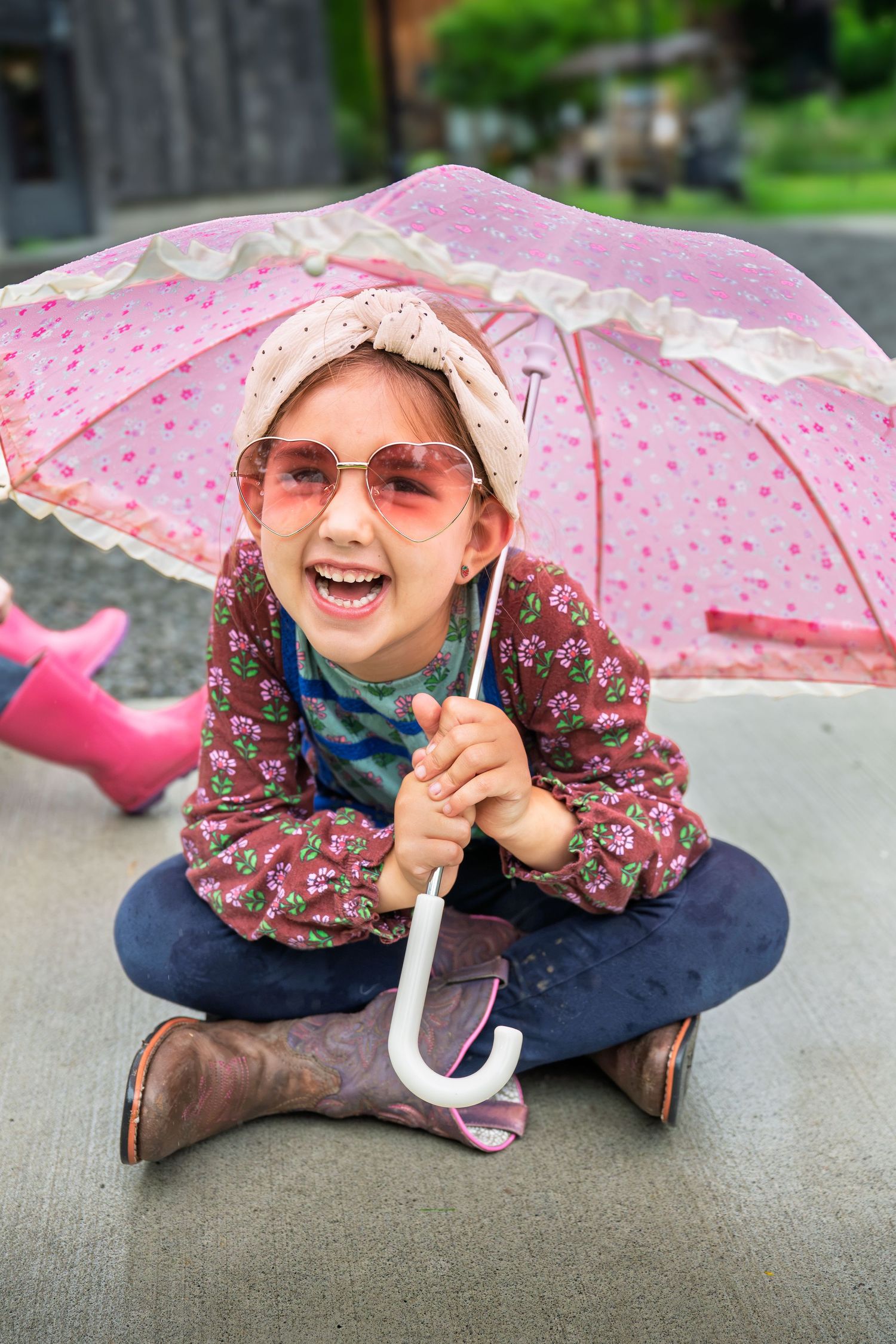 A cheerful young girl sits cross-legged holding a pink umbrella, wearing heart sunglasses and rain boots.