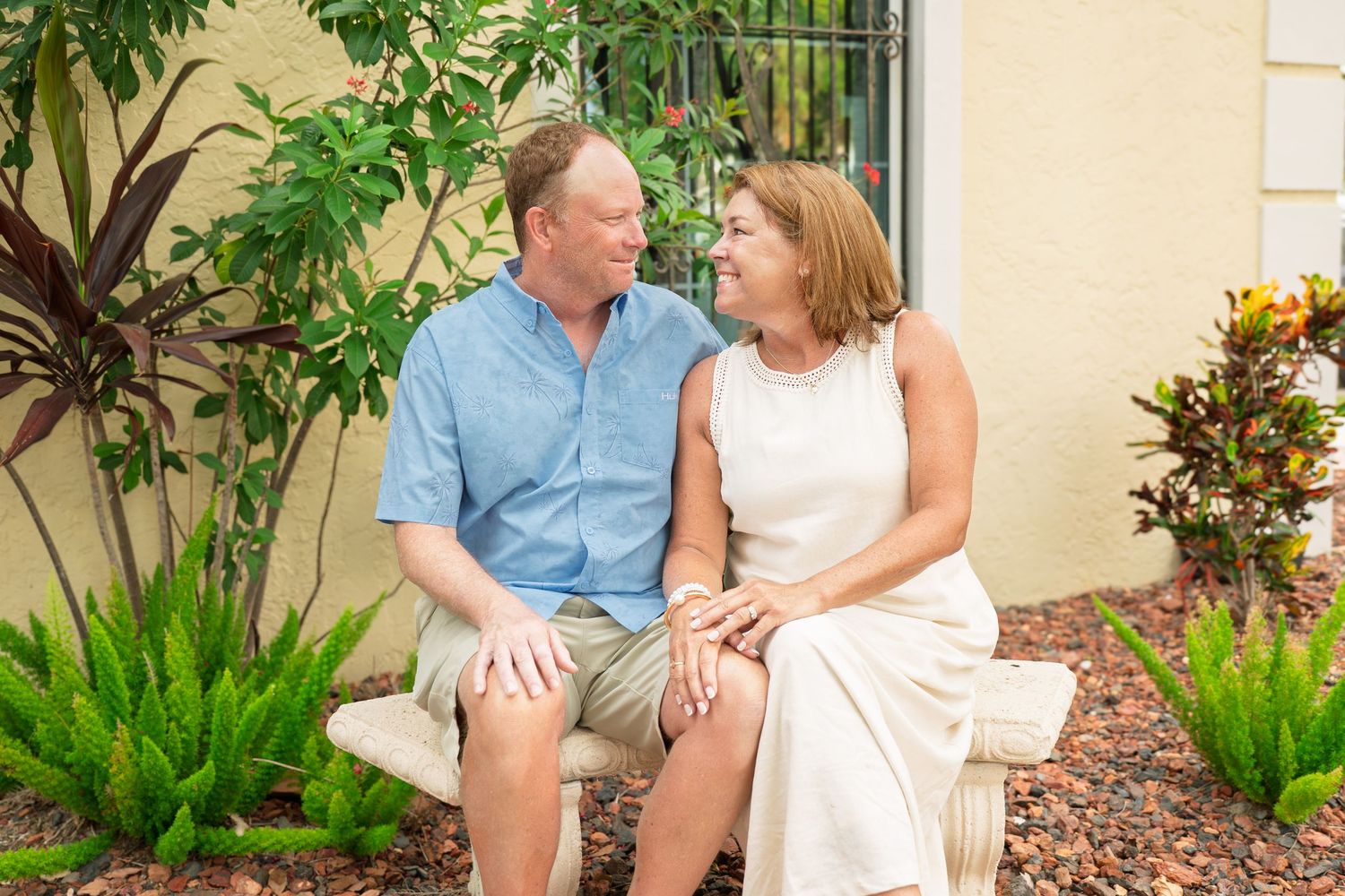 A smiling middle-aged couple sits together on a bench surrounded by lush tropical plants near a yellow building.