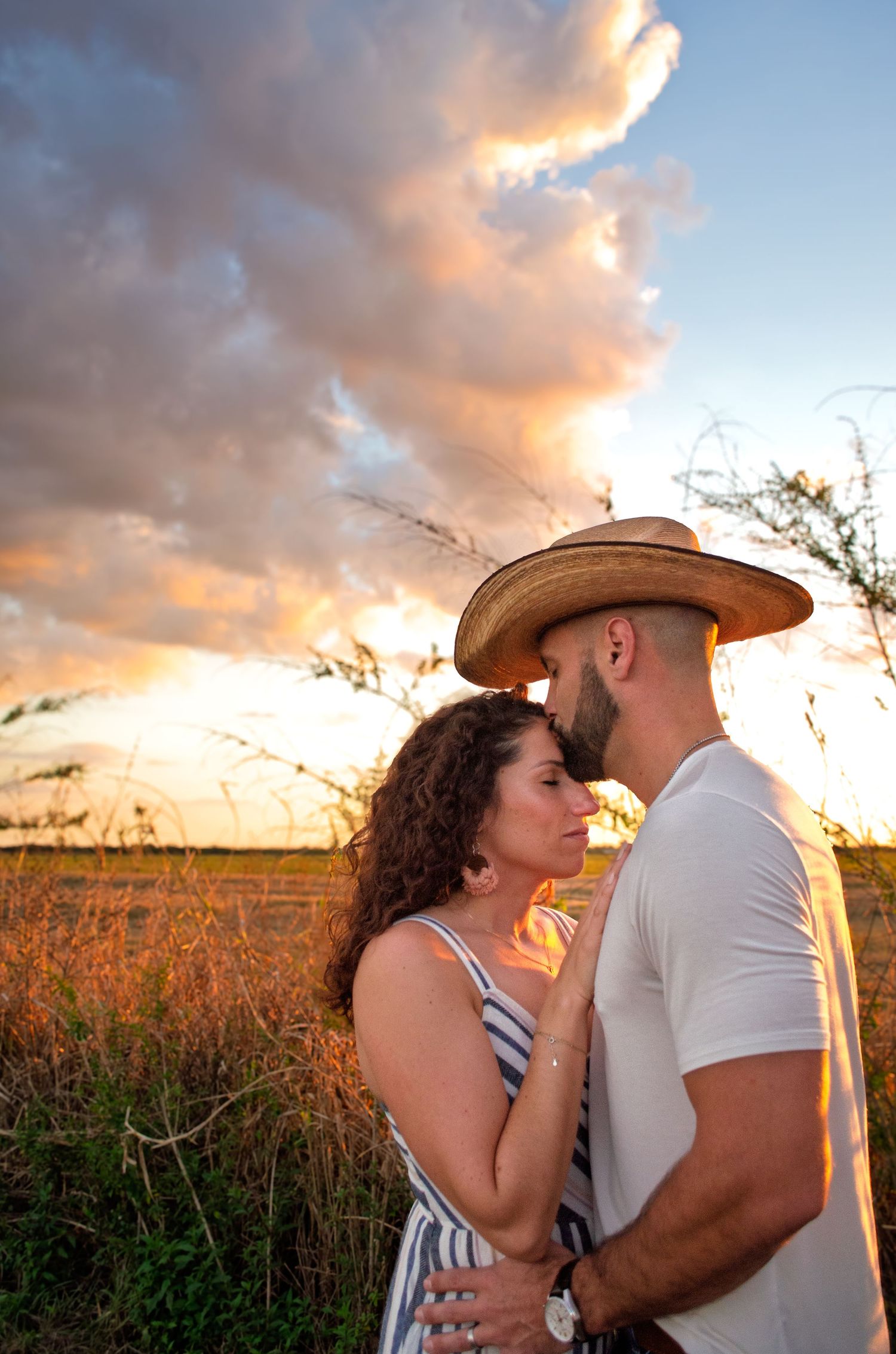 A romantic couple embraces in a golden wheat field at sunset while wearing cowboy hats and western attire.