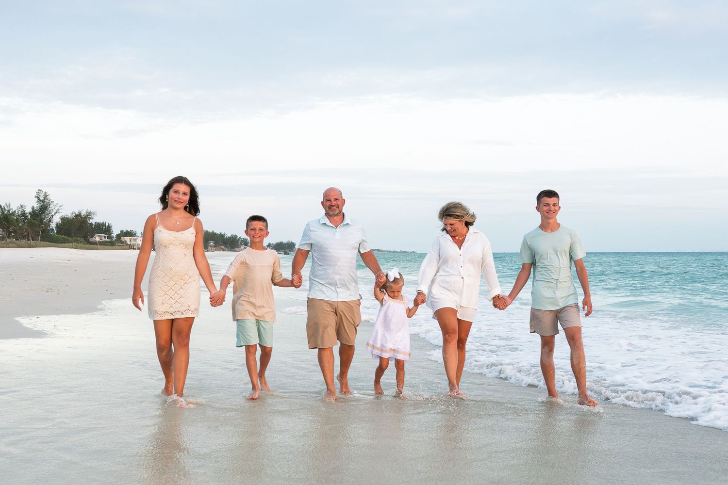 Multi-generational family of six walks hand-in-hand along a sunny sandy beach with gentle ocean waves.