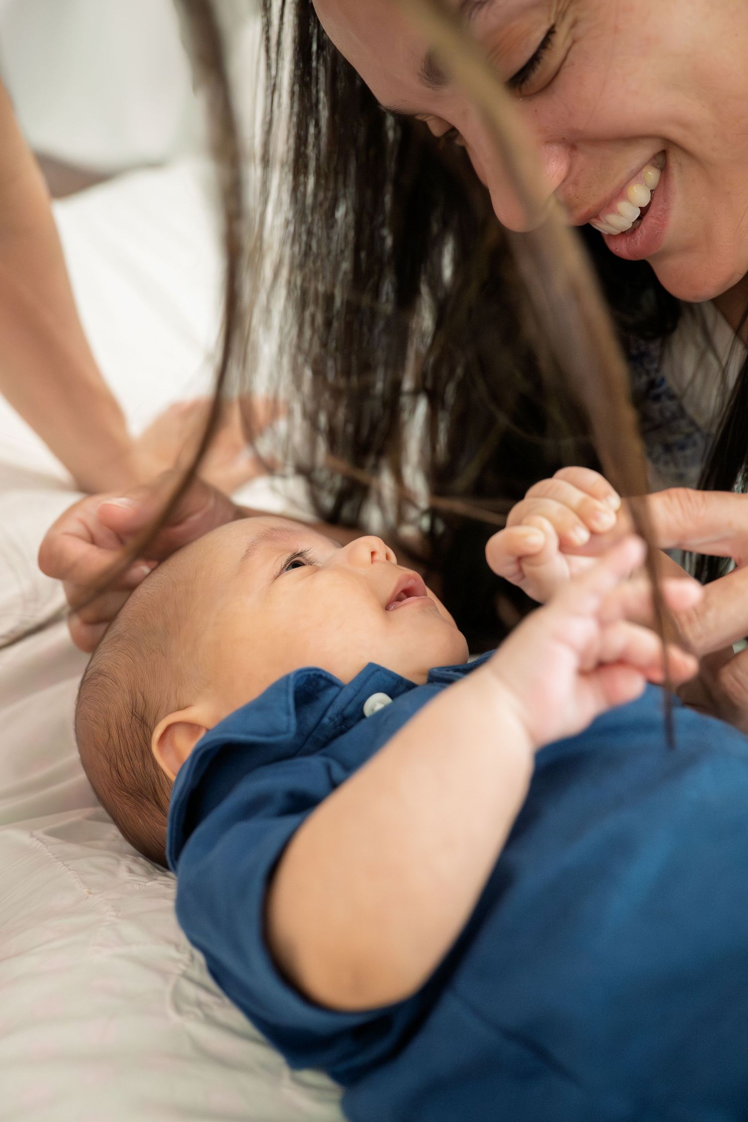 Smiling mother playing with her newborn baby dressed in blue, lying on a white bed.