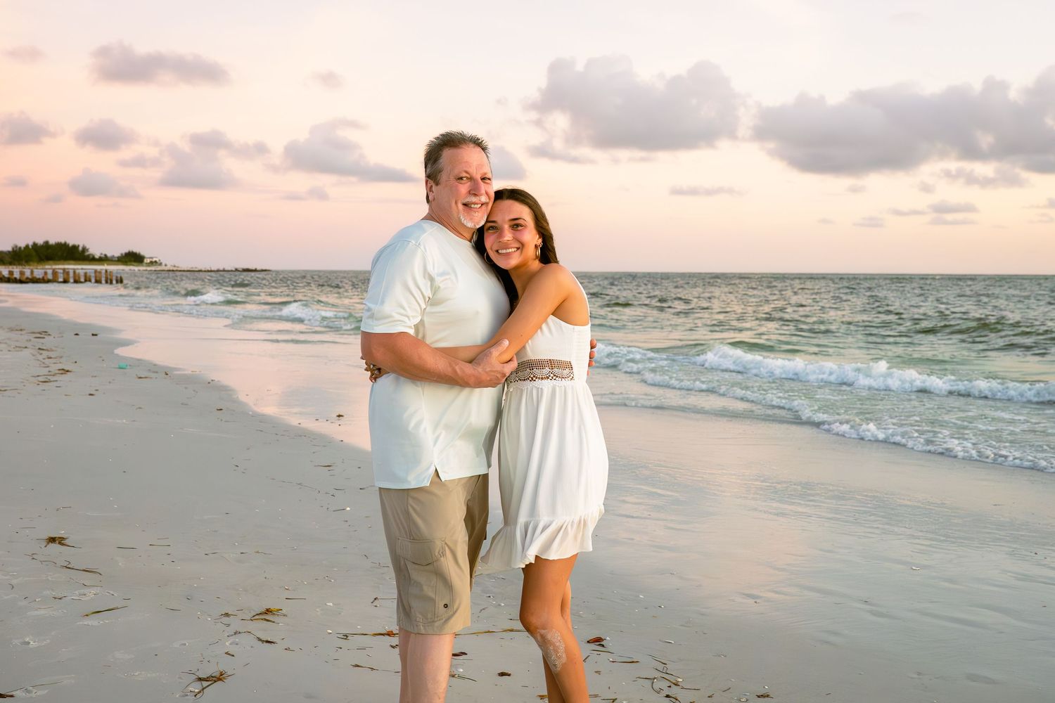 Couple embracing and smiling on a sandy beach at sunset with gentle waves and pastel sky behind them.