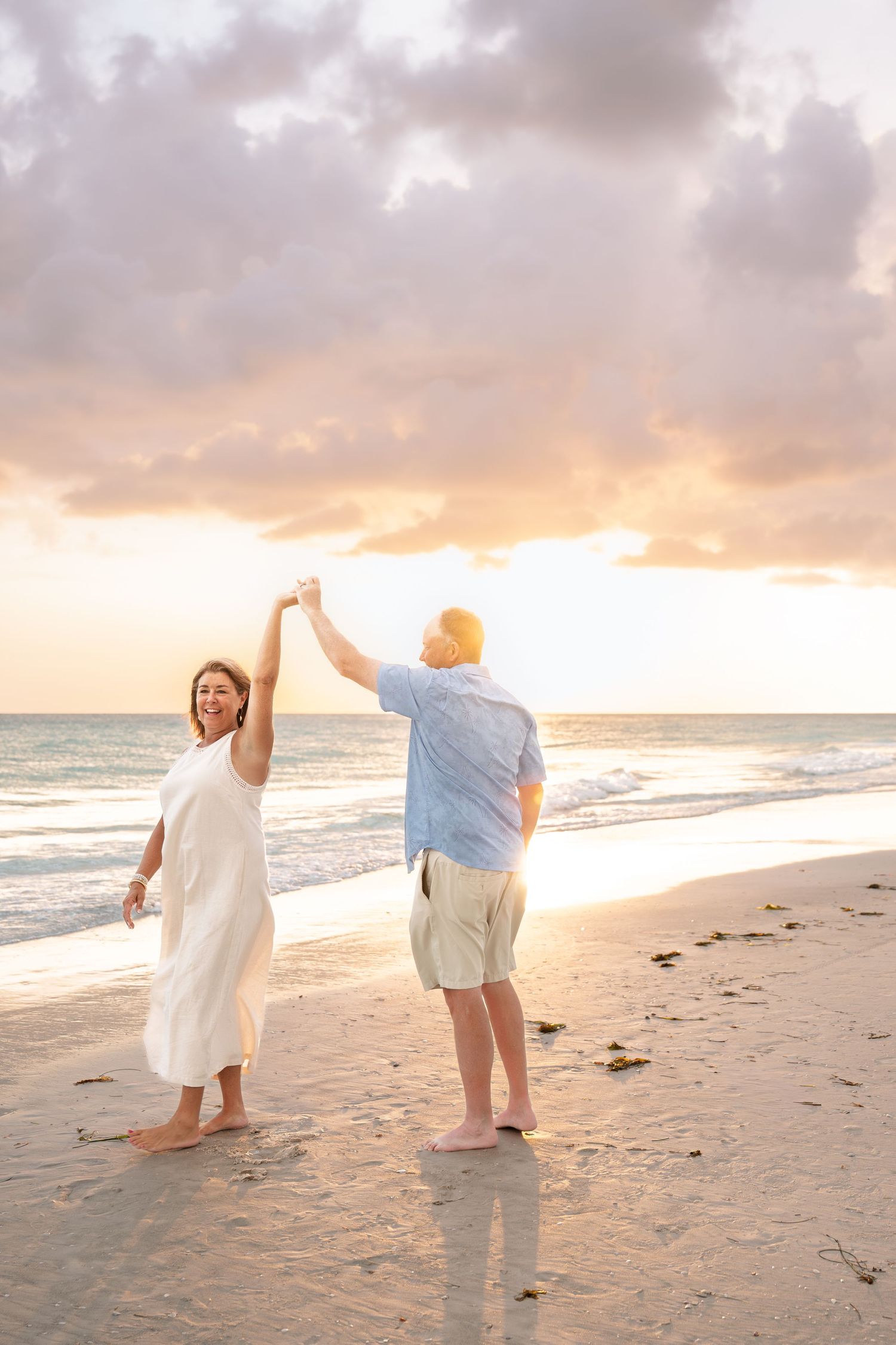 Couple dancing on beach at sunset, woman in white dress being twirled by man in blue shirt, ocean waves in background.