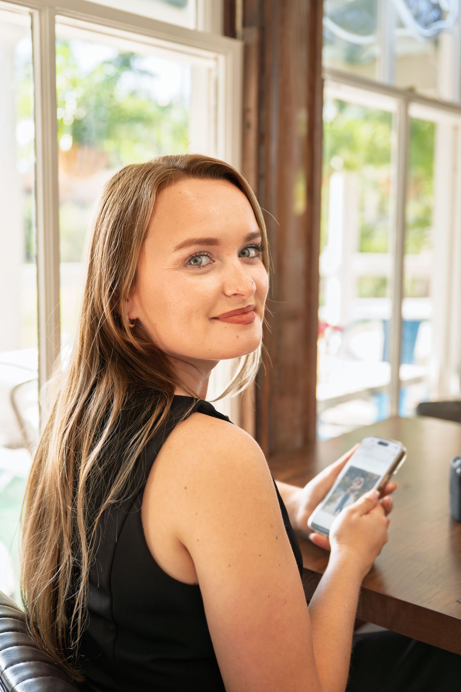 Young woman with long brown hair smiling over shoulder while holding smartphone at cafe table near bright windows.