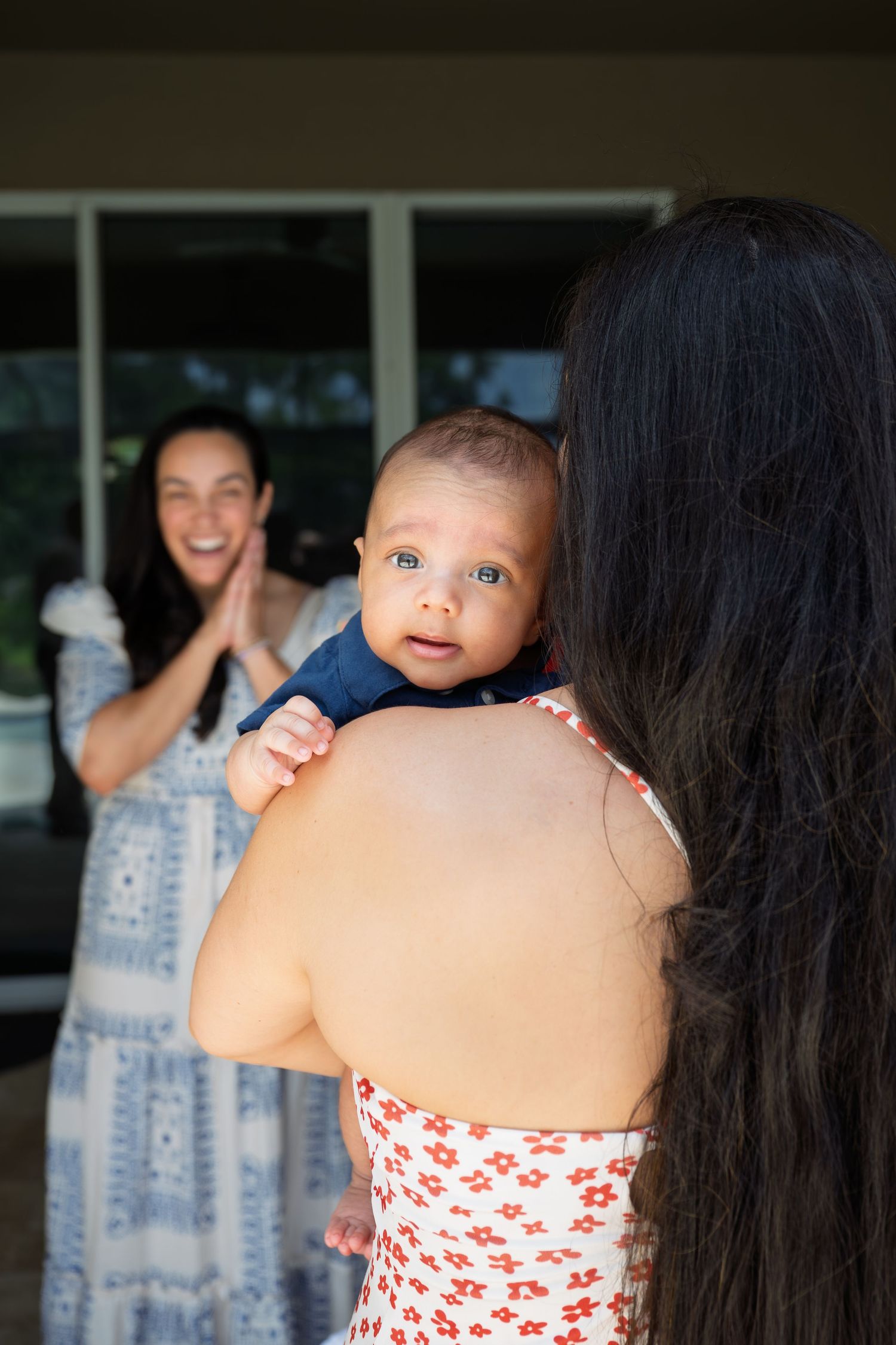 A mother holds her baby over her shoulder while an excited woman claps her hands in the background.