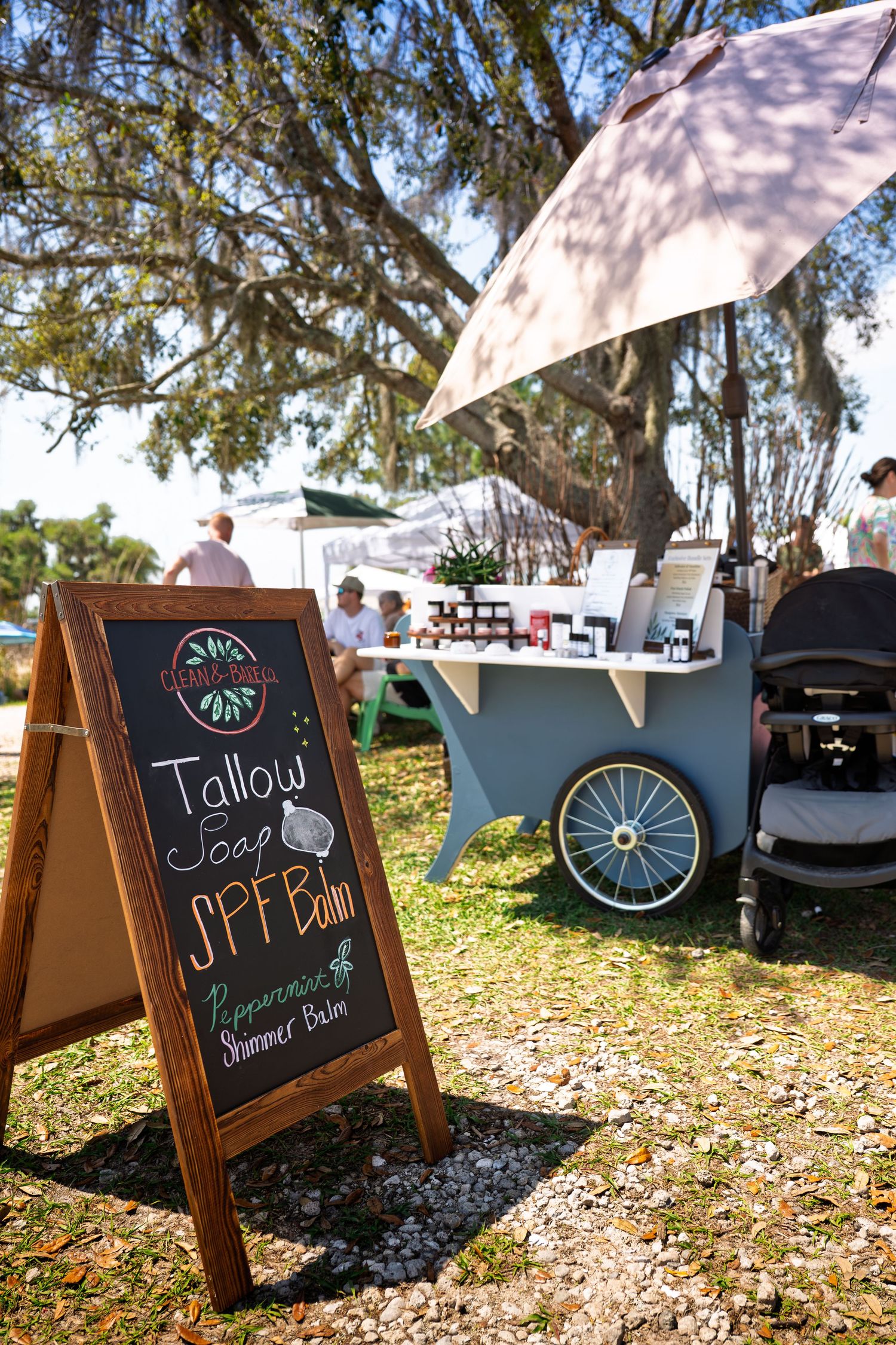 Outdoor farmers market booth with chalkboard sign advertising tallow soap and SPF balm under a large oak tree.