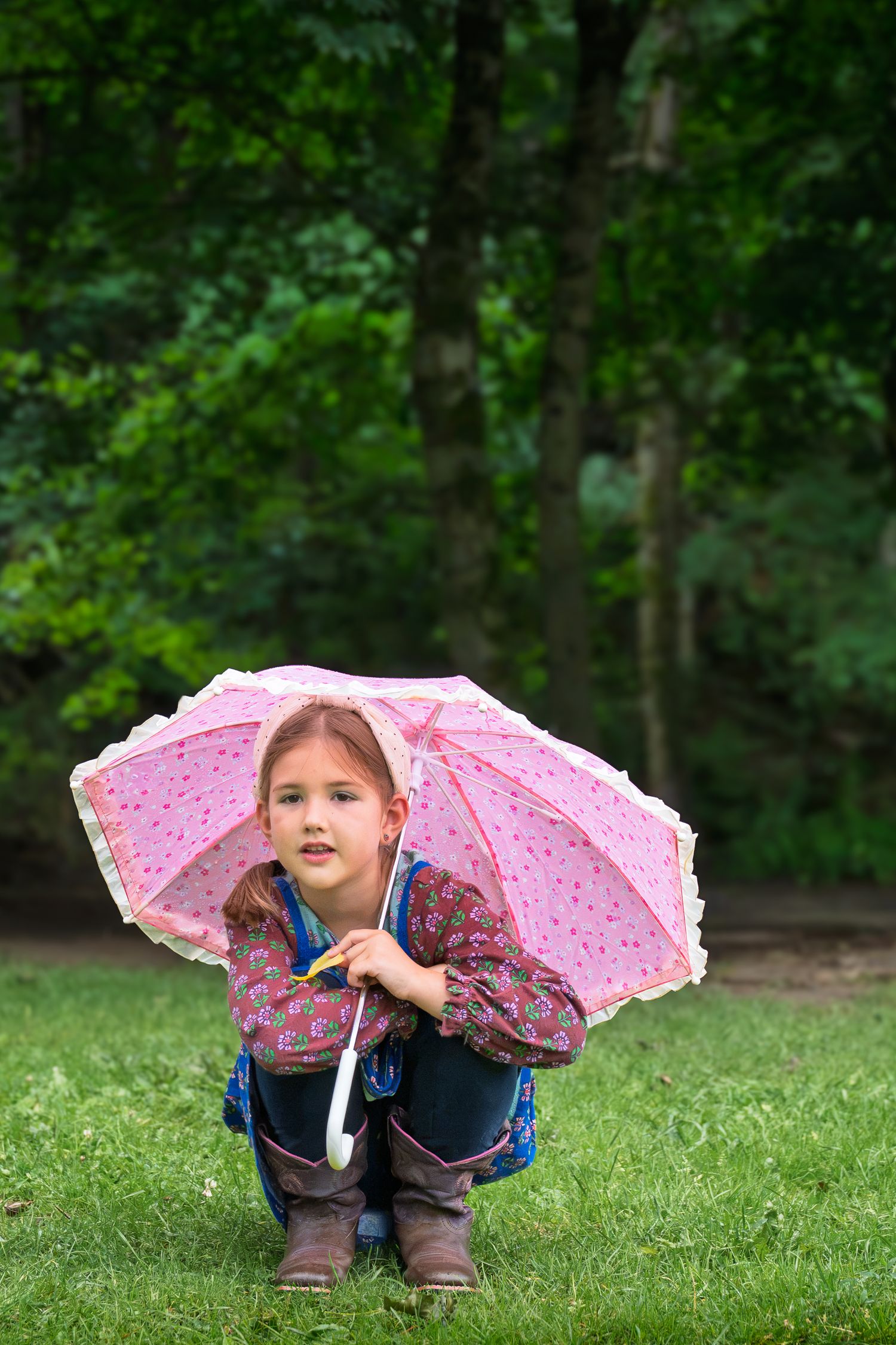 Young girl crouching on grass holding a pink umbrella, wearing boots and a colorful jacket in a green park.