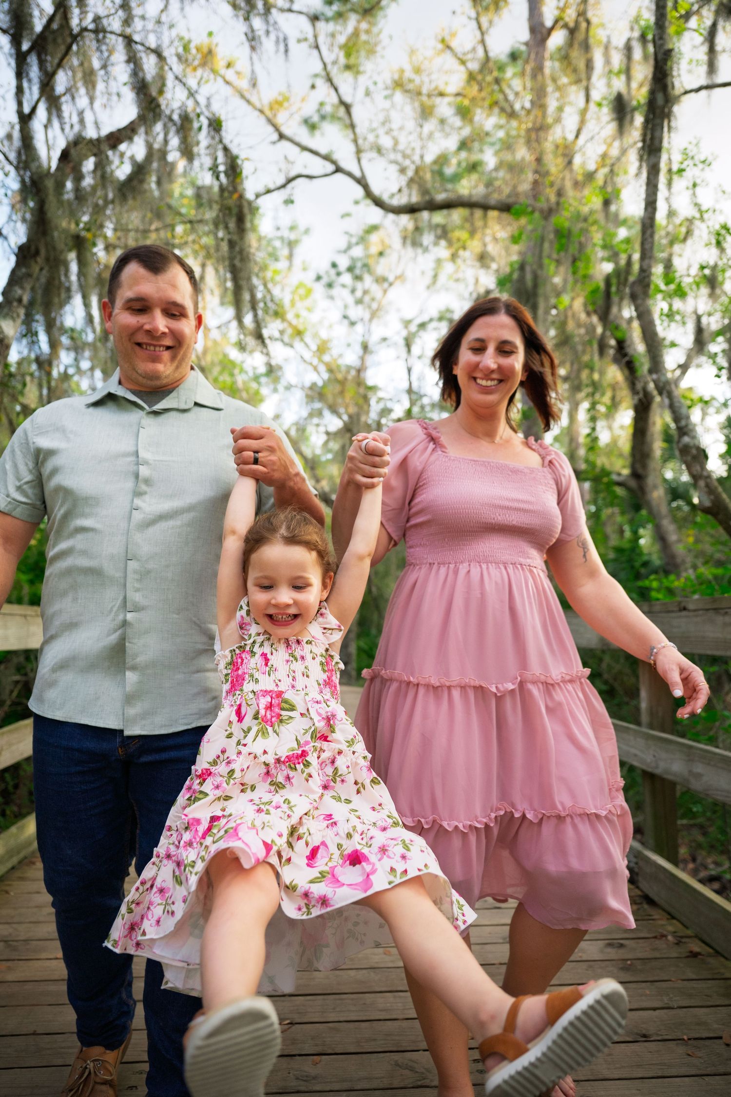 A family poses playfully on a wooden boardwalk surrounded by Spanish moss and trees in a natural outdoor setting.
